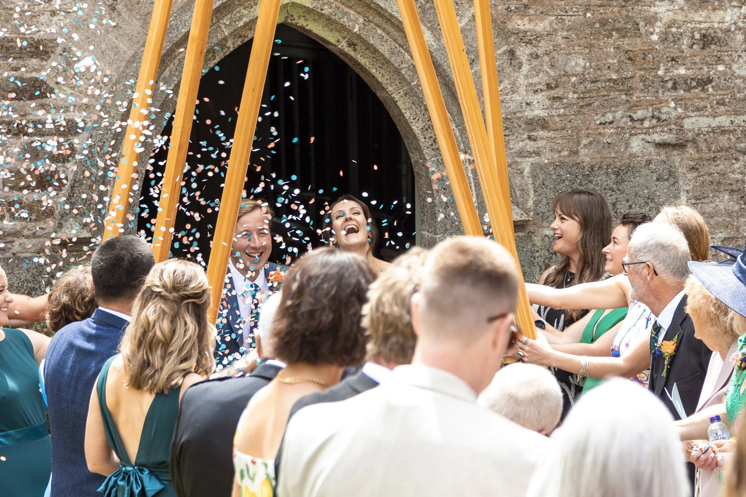 People celebrating outside with confetti falling, some holding a wooden arch, in front of a brick wall, during a joyful event.
