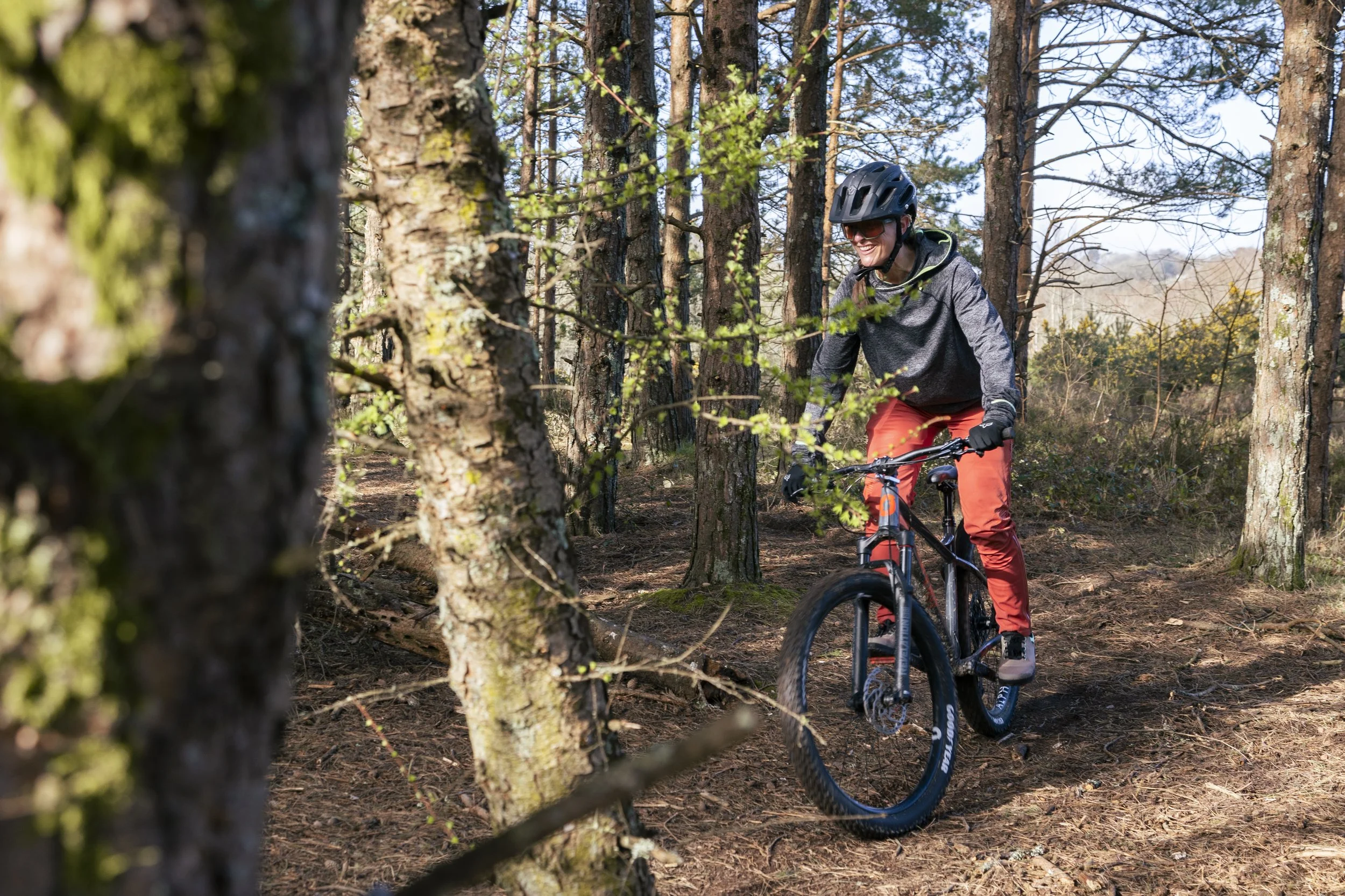 A person riding a mountain bike on a forest trail, wearing a helmet and outdoor clothing, smiling as they navigate through trees.