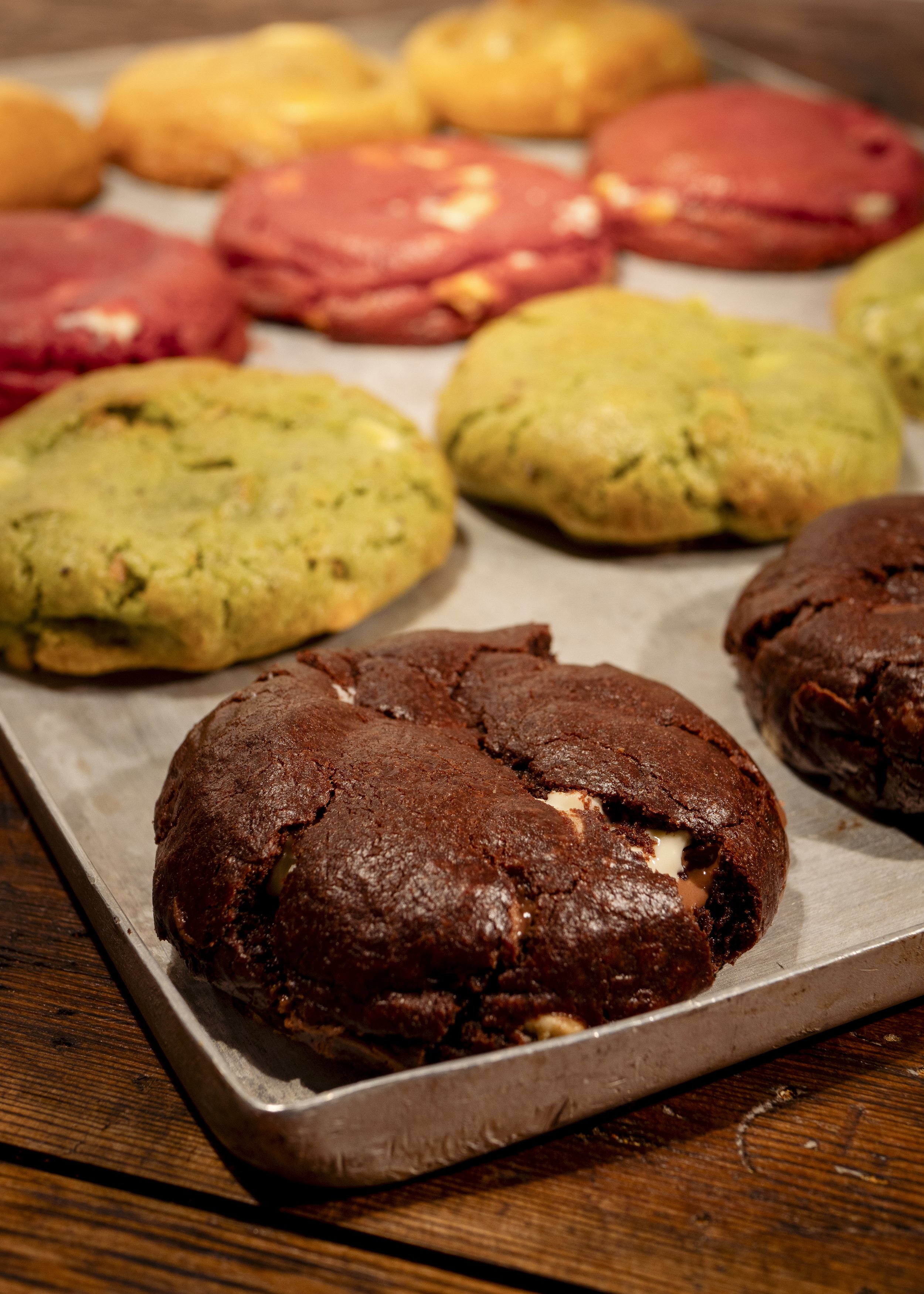 An assortment of freshly baked cookies with different colors and textures on a baking sheet, including chocolate, green, red, and yellow cookies, on a wooden surface.