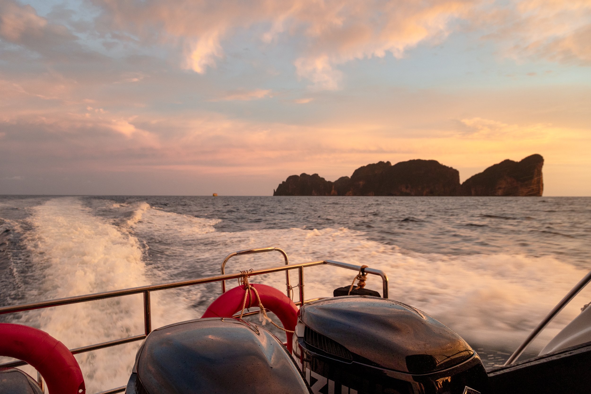 View from a boat at sunset, with motor engines at the foreground, a wake trail in the water, and an island in the distance under a pink and orange sky.