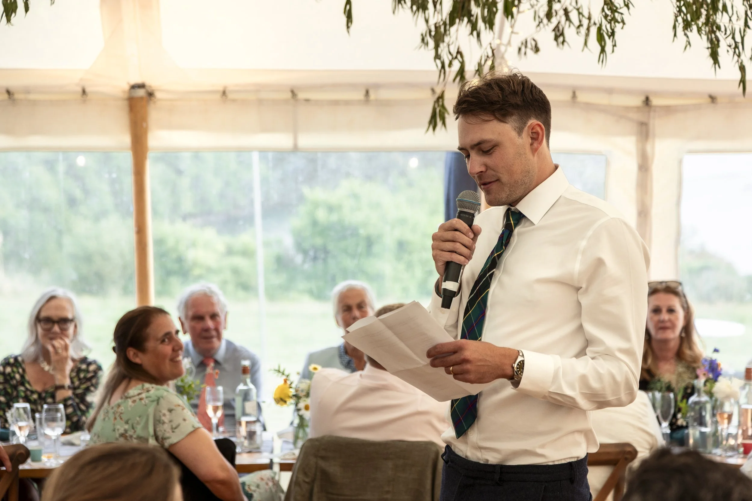 A man in a white shirt and striped tie giving a speech while holding a microphone and reading from notes during a social gathering at a decorated outdoor tent, with seated guests and greenery in the background.
