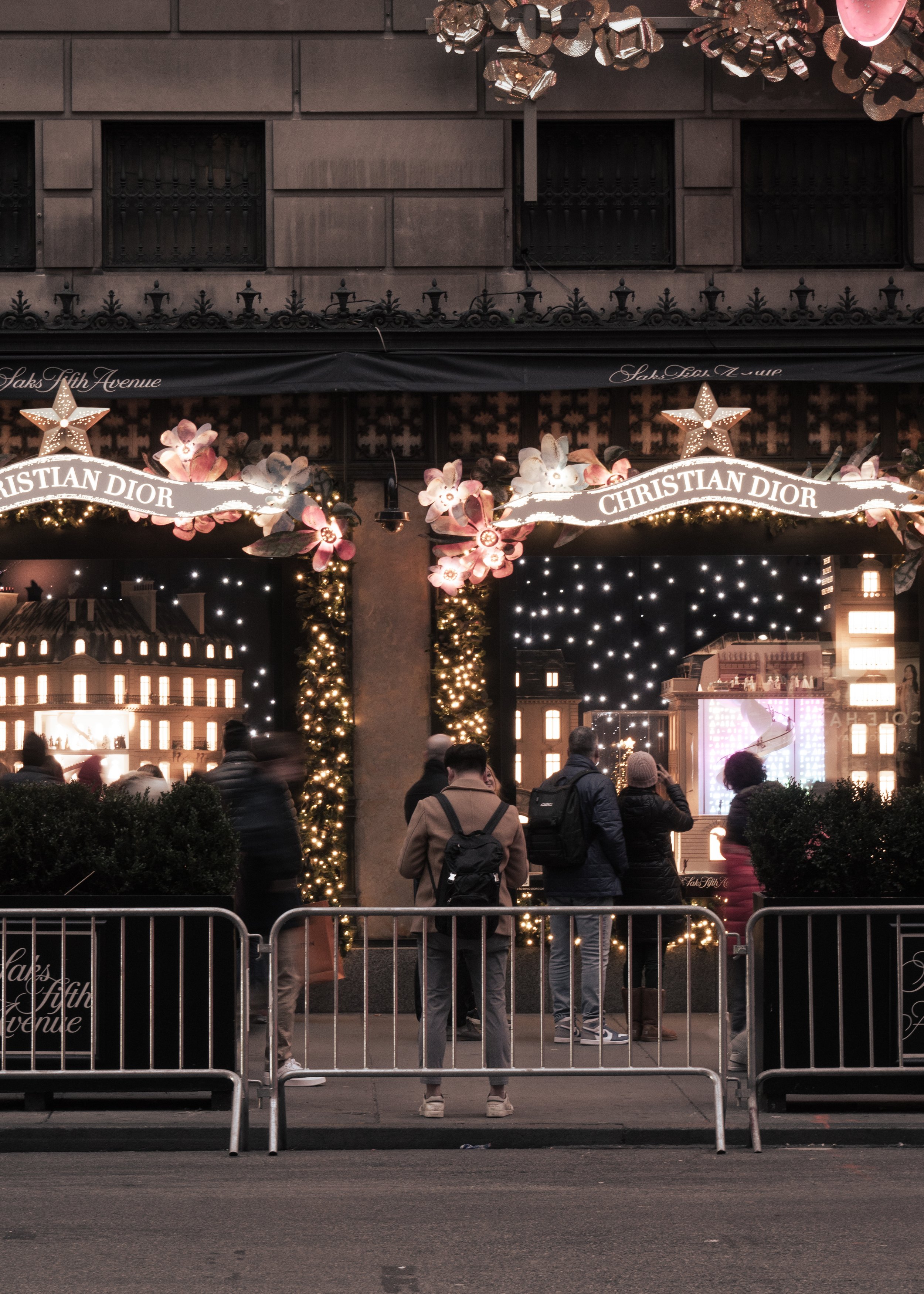 People standing behind a metal barricade in front of a festive display with illuminated buildings, Christmas trees, and Christian Dior signs, decorated with pink flowers, stars, and string lights.