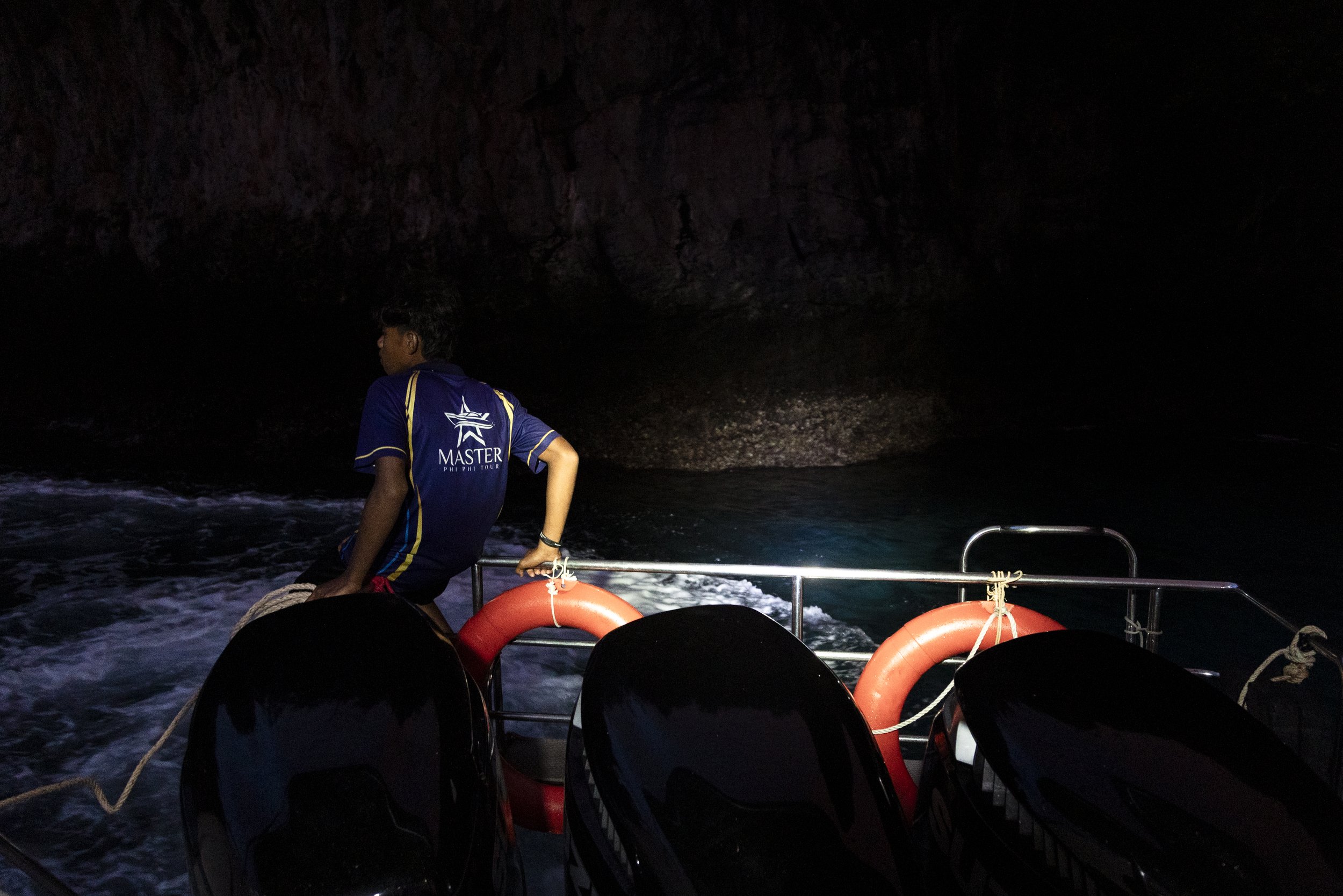 A person on a boat at night near a dark rocky shoreline, wearing a blue shirt with a logo, standing near the lifebuoys and motor engines.