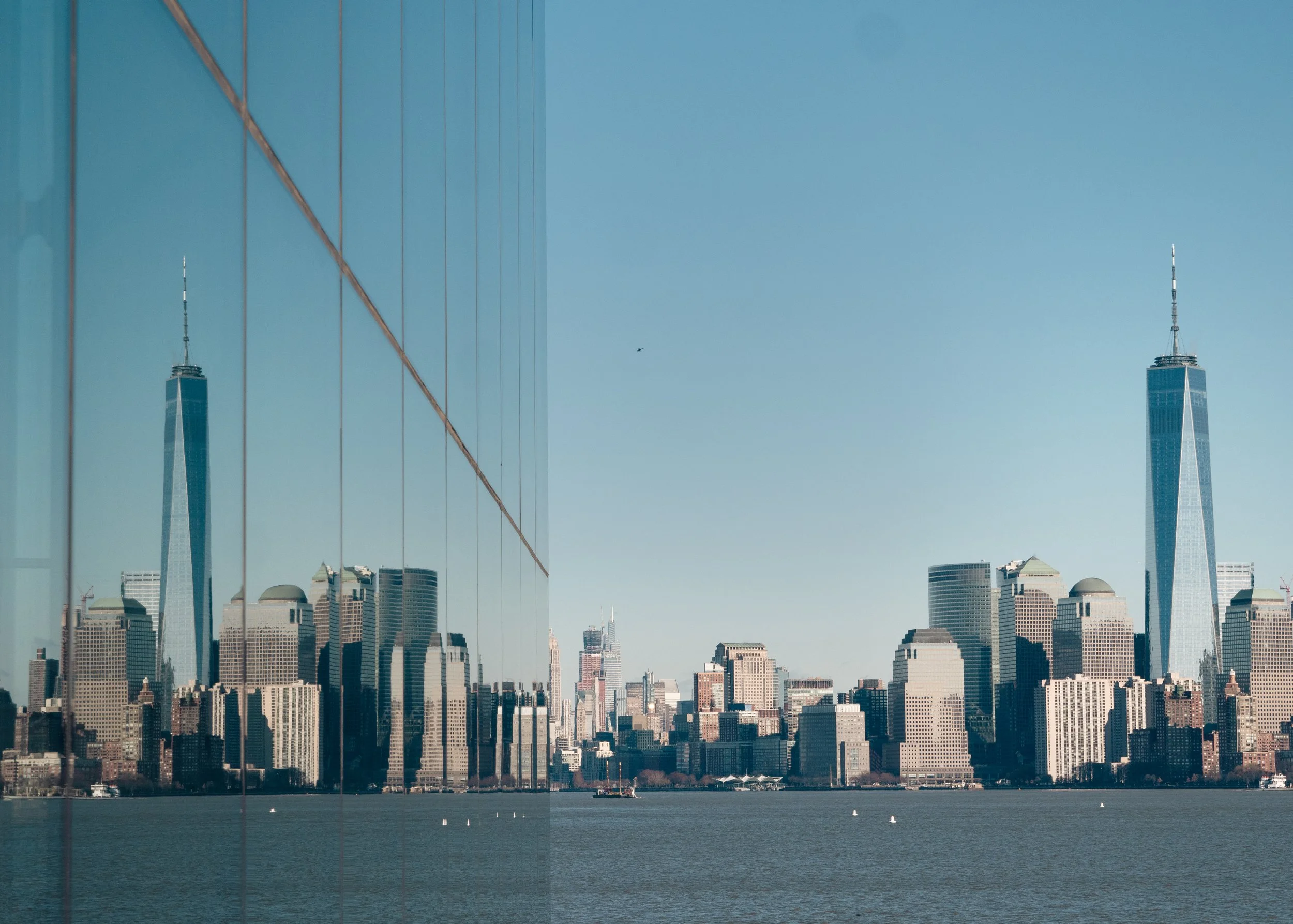 View of Manhattan skyline with One World Trade Center and other buildings, reflected on the glass facade of a nearby building, with a body of water in the foreground and a clear blue sky.