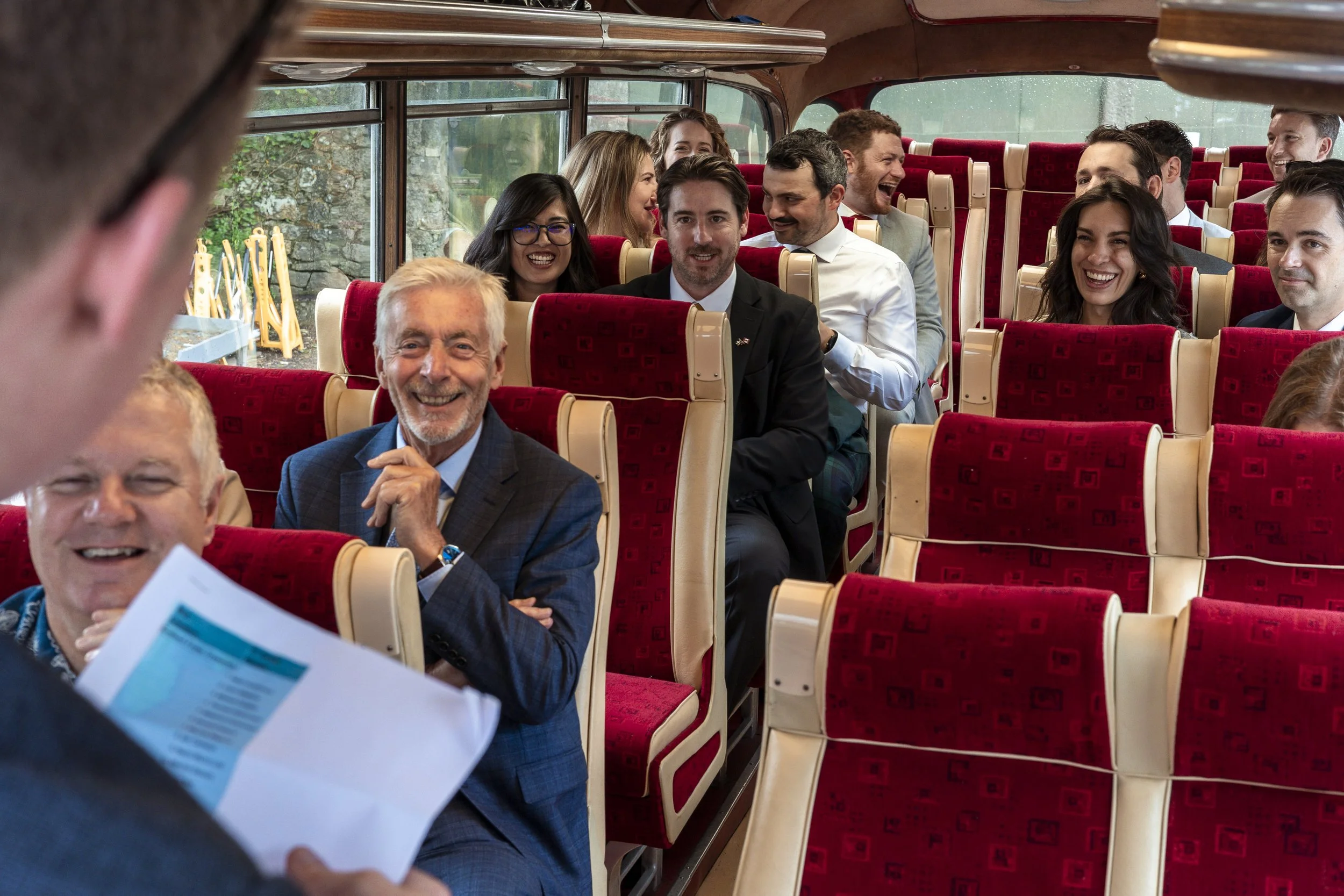 Group of people sitting on a bus, smiling and laughing during a presentation or speech, with a person holding papers in the foreground.