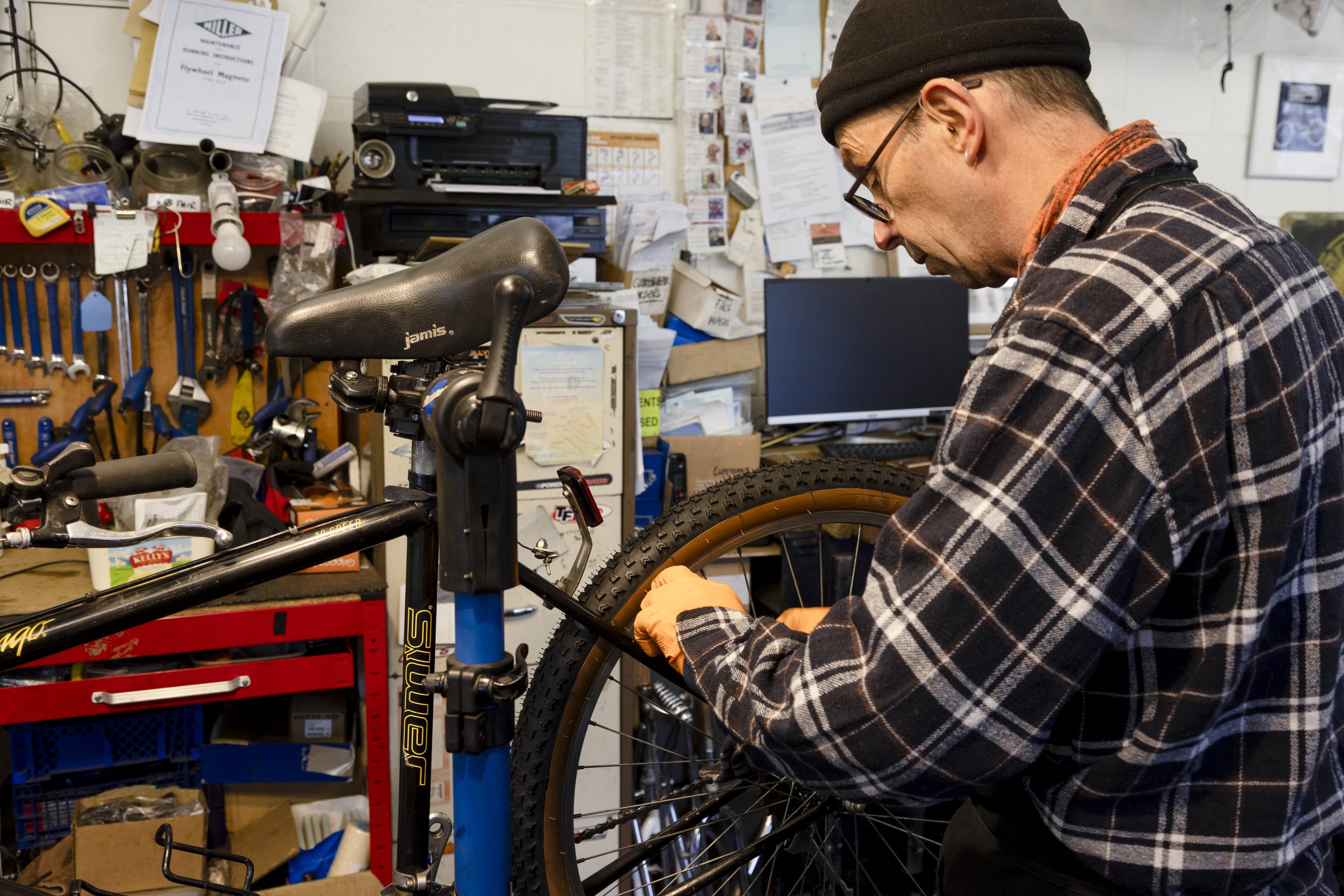A man wearing glasses, a black beanie, and a plaid shirt working on a bicycle in a cluttered workshop.