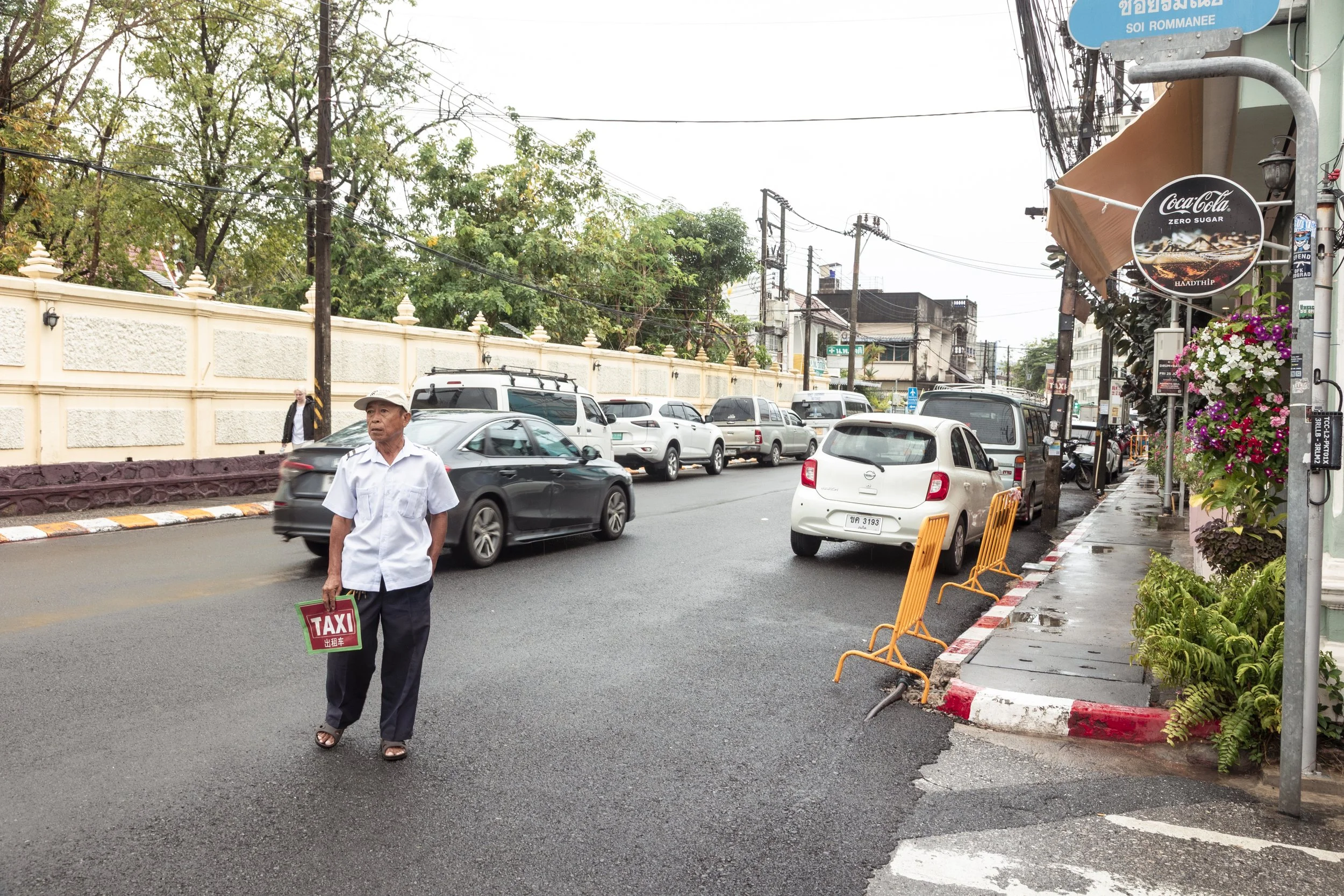 A man holding a small taxi sign stands on a wet city street with parked cars and storefronts, including Coca-Cola signage and hanging flower baskets.