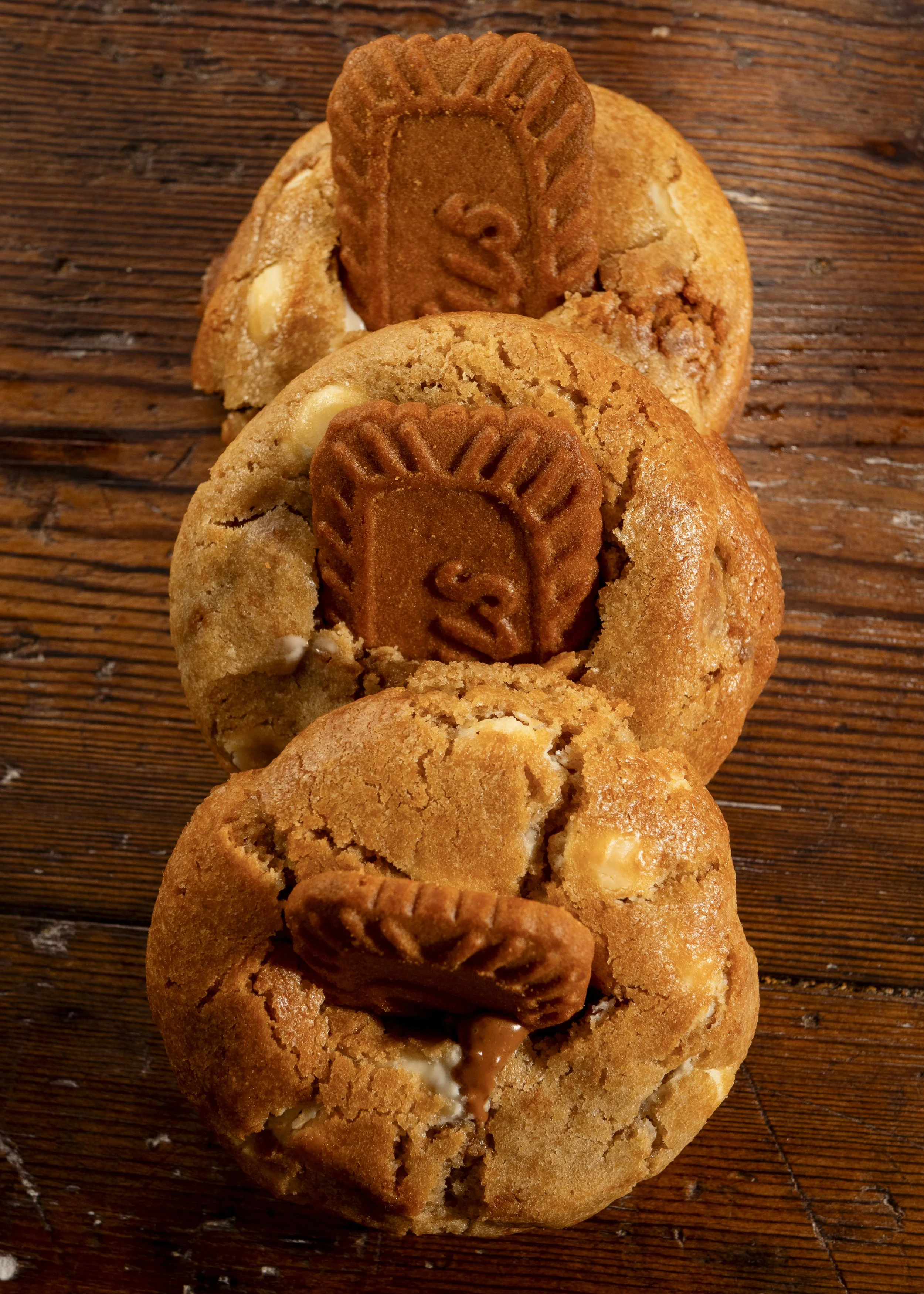 Three cookies with white chocolate chips and a Biscoff cookie on top, arranged in a vertical line on a wooden surface.