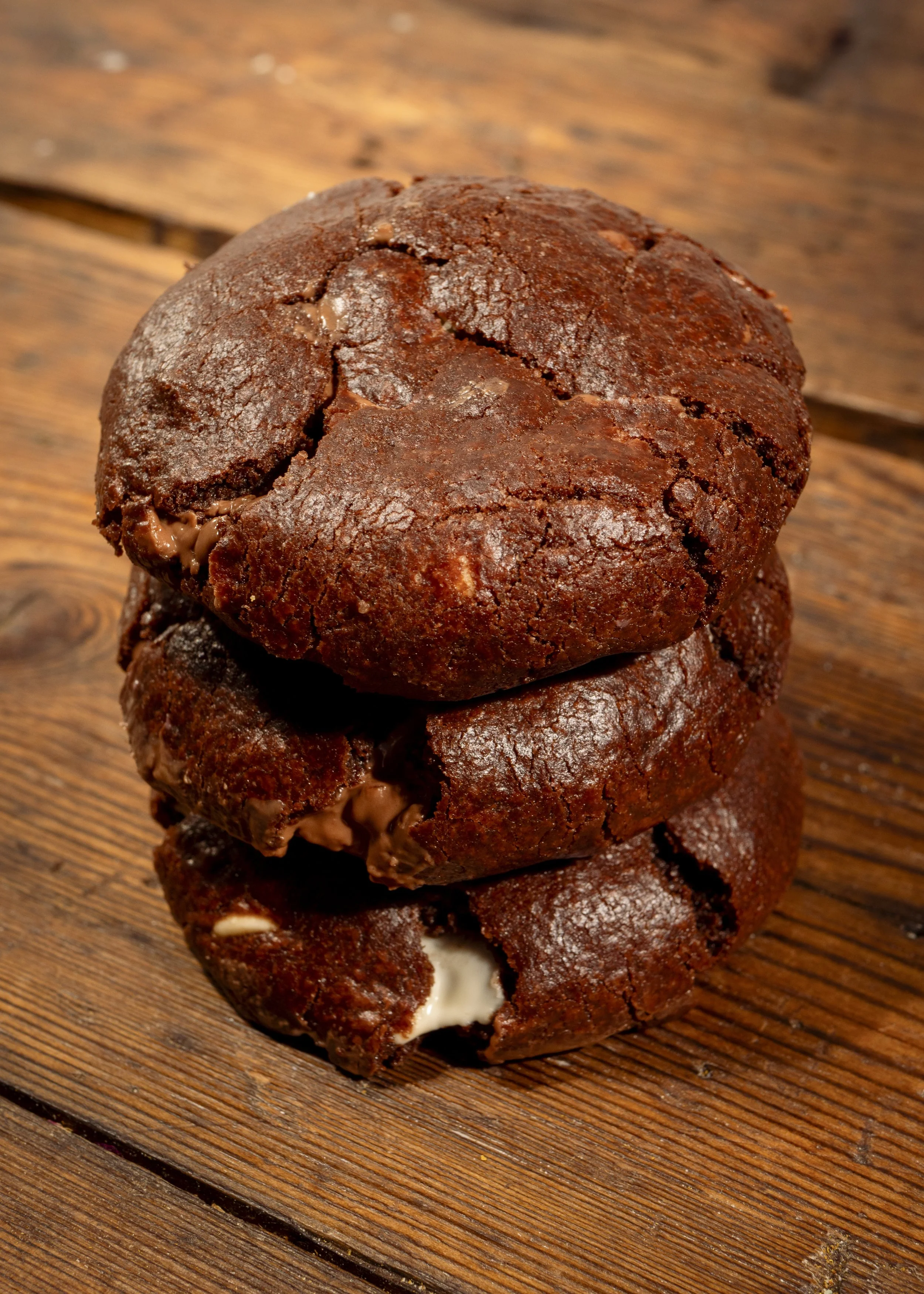 Three chocolate cookies stacked on a wooden surface, with a chewy and slightly cracked appearance.