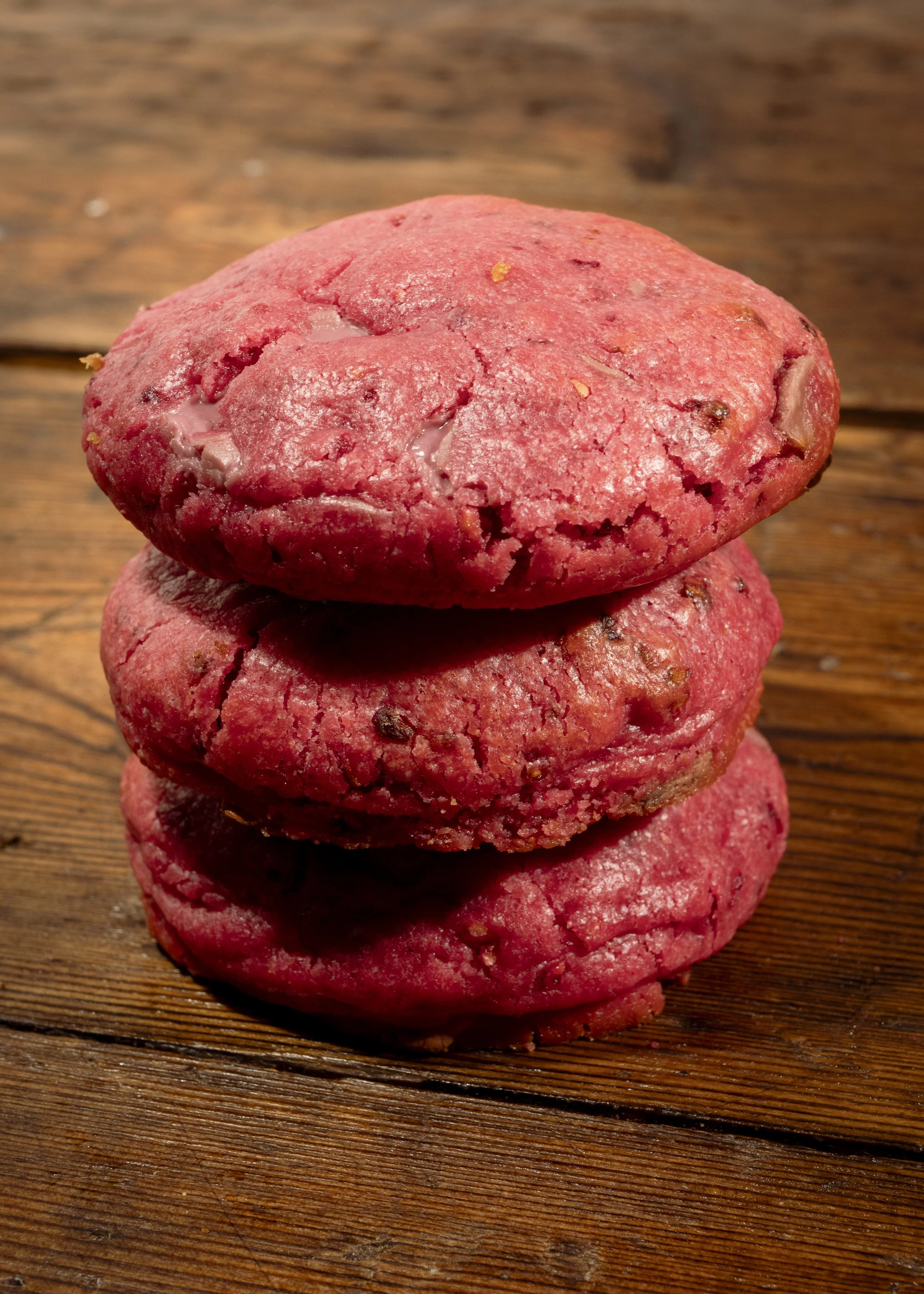 Stack of three red cookies with chocolate chips on a wooden surface.