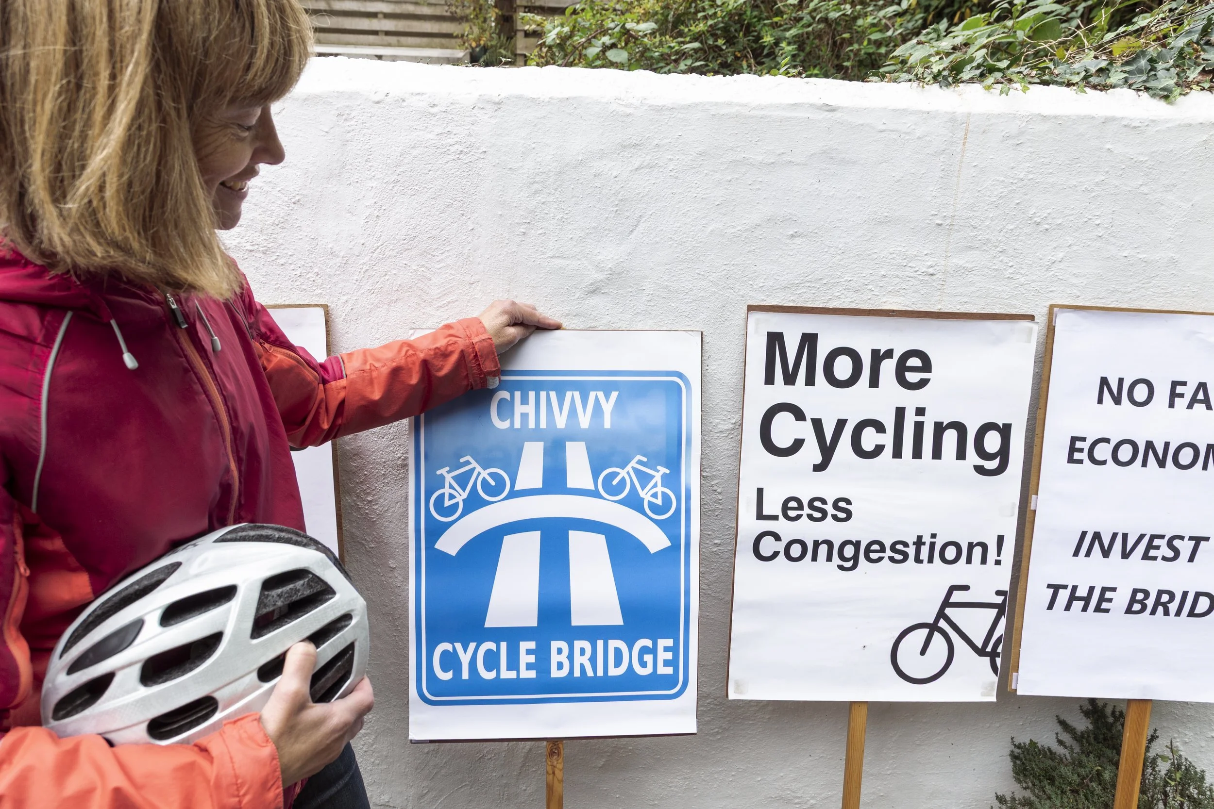 A woman in a pink jacket holding a white bicycle helmet, standing next to a blue sign that says "Cycle Bridge" with a graphic of a bike and a bridge. There are other signs with messages promoting more cycling and less congestion, and a bike graphic.