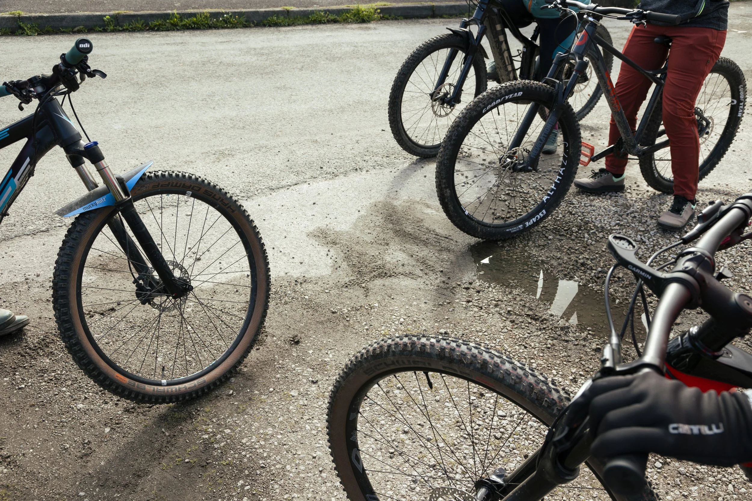 Group of people with mountain bikes gathered outdoors on a gravel path, with some standing and their bikes parked around them.