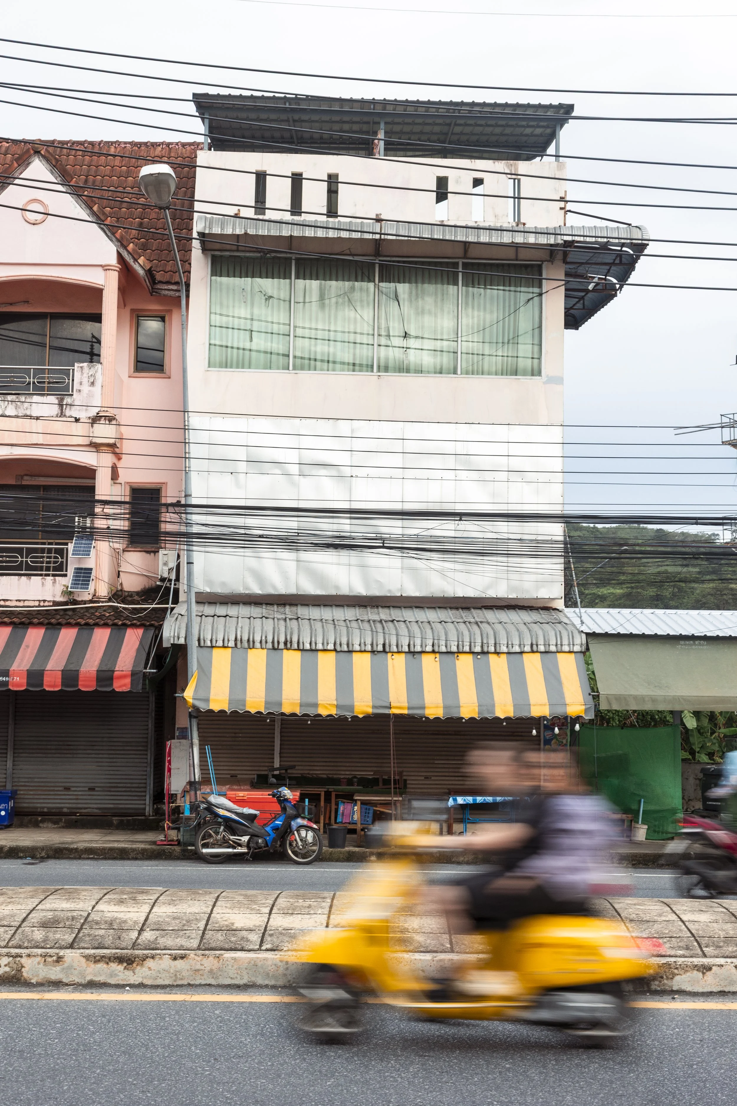 A street scene showing the front of a multi-story building with an awning, motorcycles parked in front, and a person riding a yellow scooter blurred in motion.