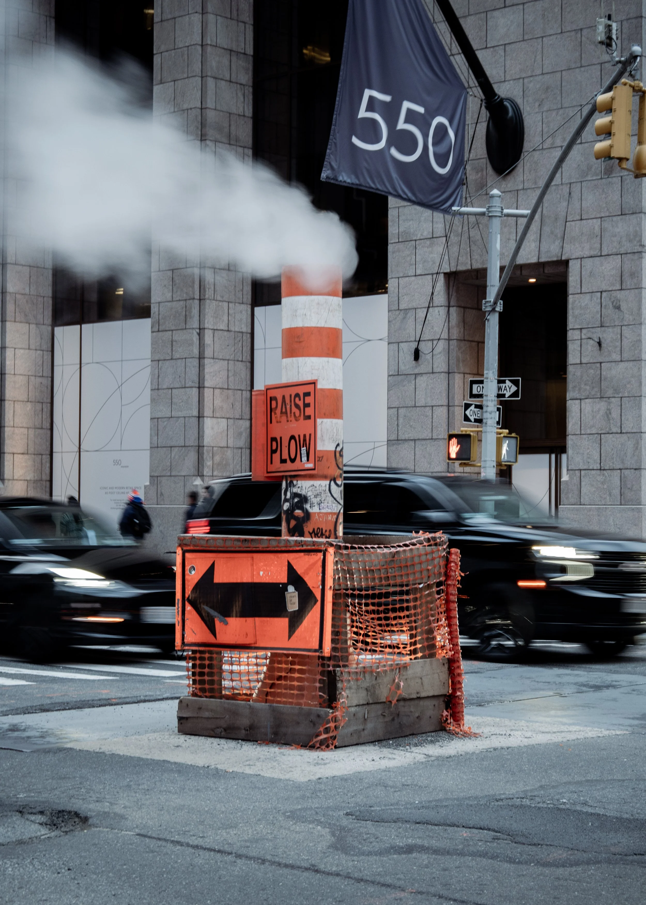 Street construction site with orange barrier, 'Raise Plow' sign, and black arrow sign indicating left or right, in front of a building marked '550'. Cars are passing by and a street light and traffic signals are visible.