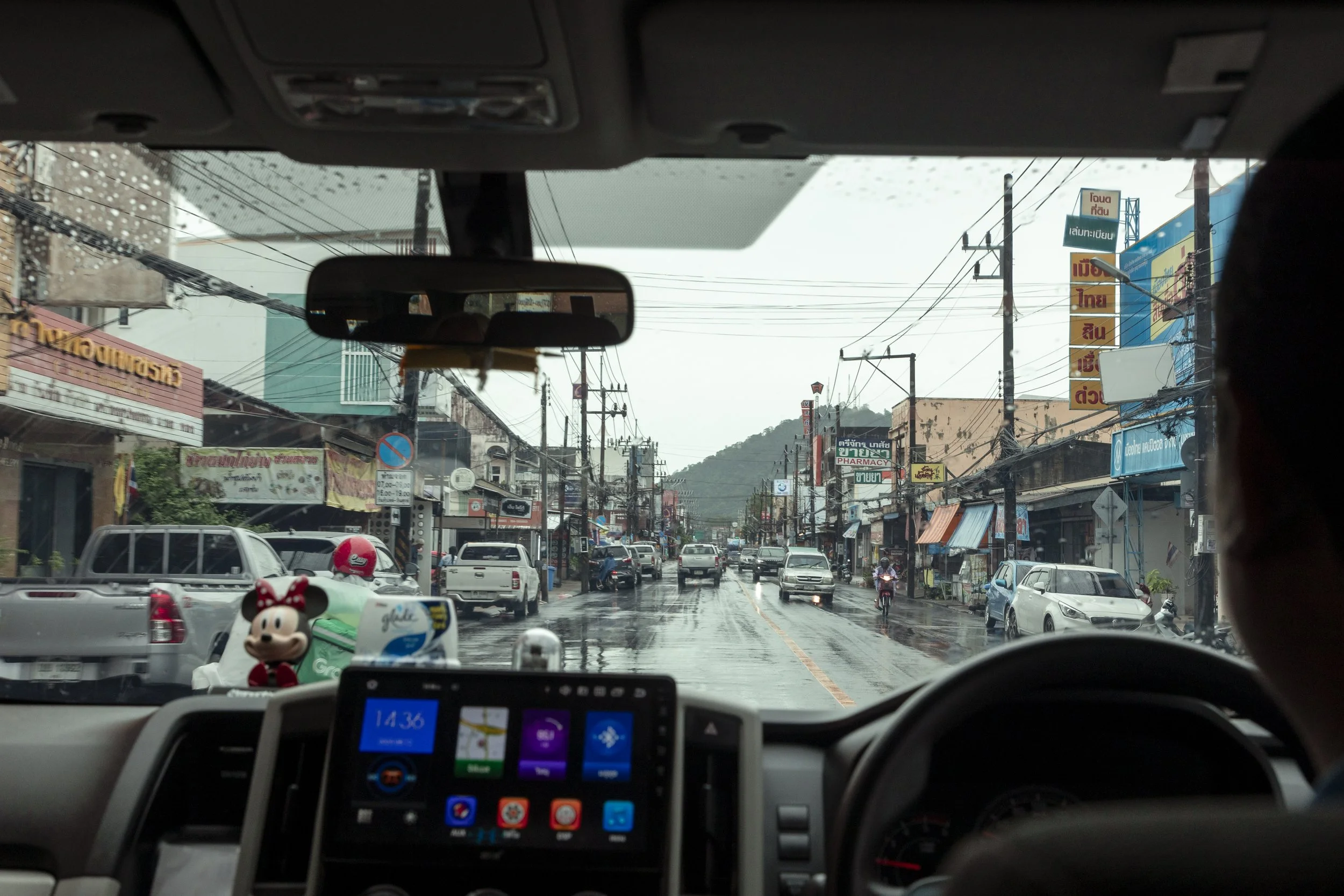 View from inside a car on a rainy street in a busy town in Thailand, with storefronts, vehicles, and electrical wires visible through the rain-streaked windshield.