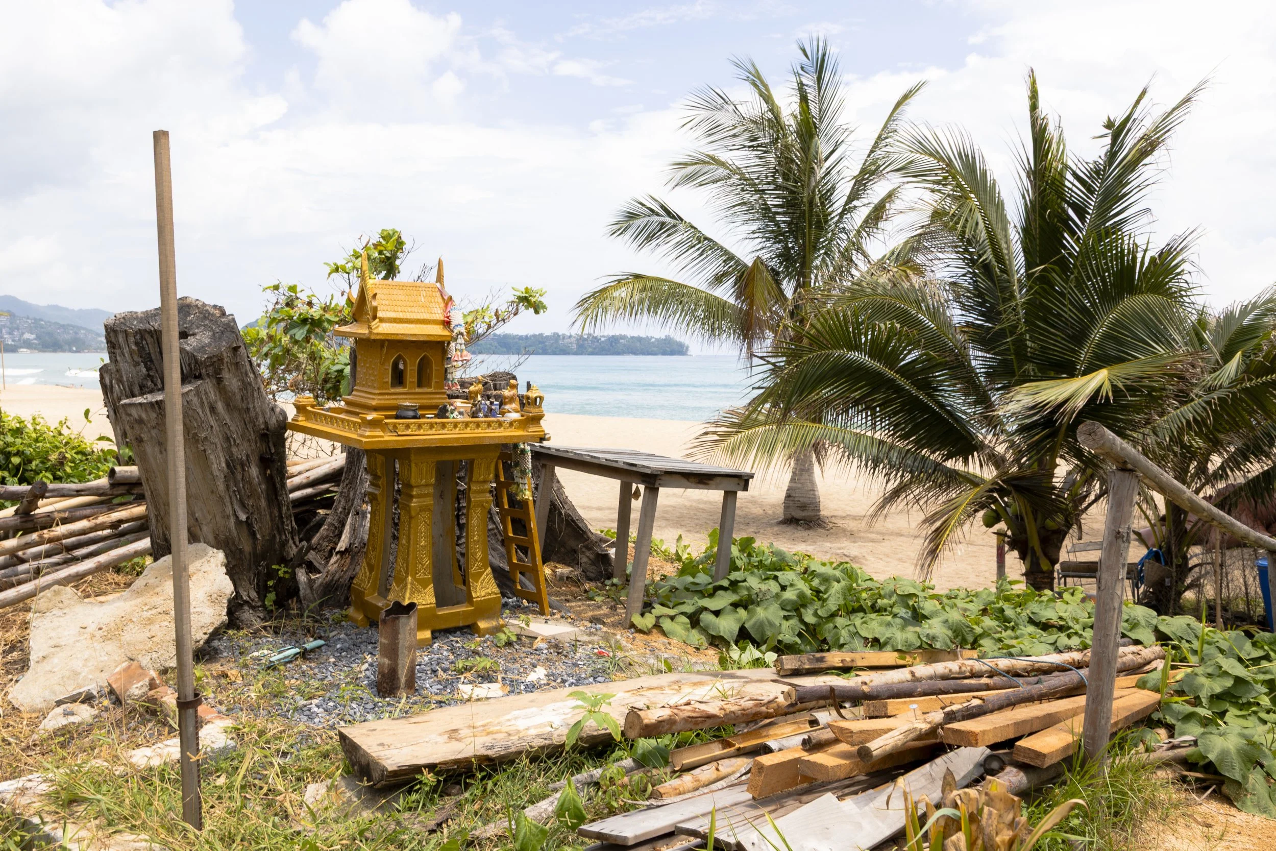 A small shrine on a beach with palm trees, a wooden table, and piles of wood and rocks in the foreground.