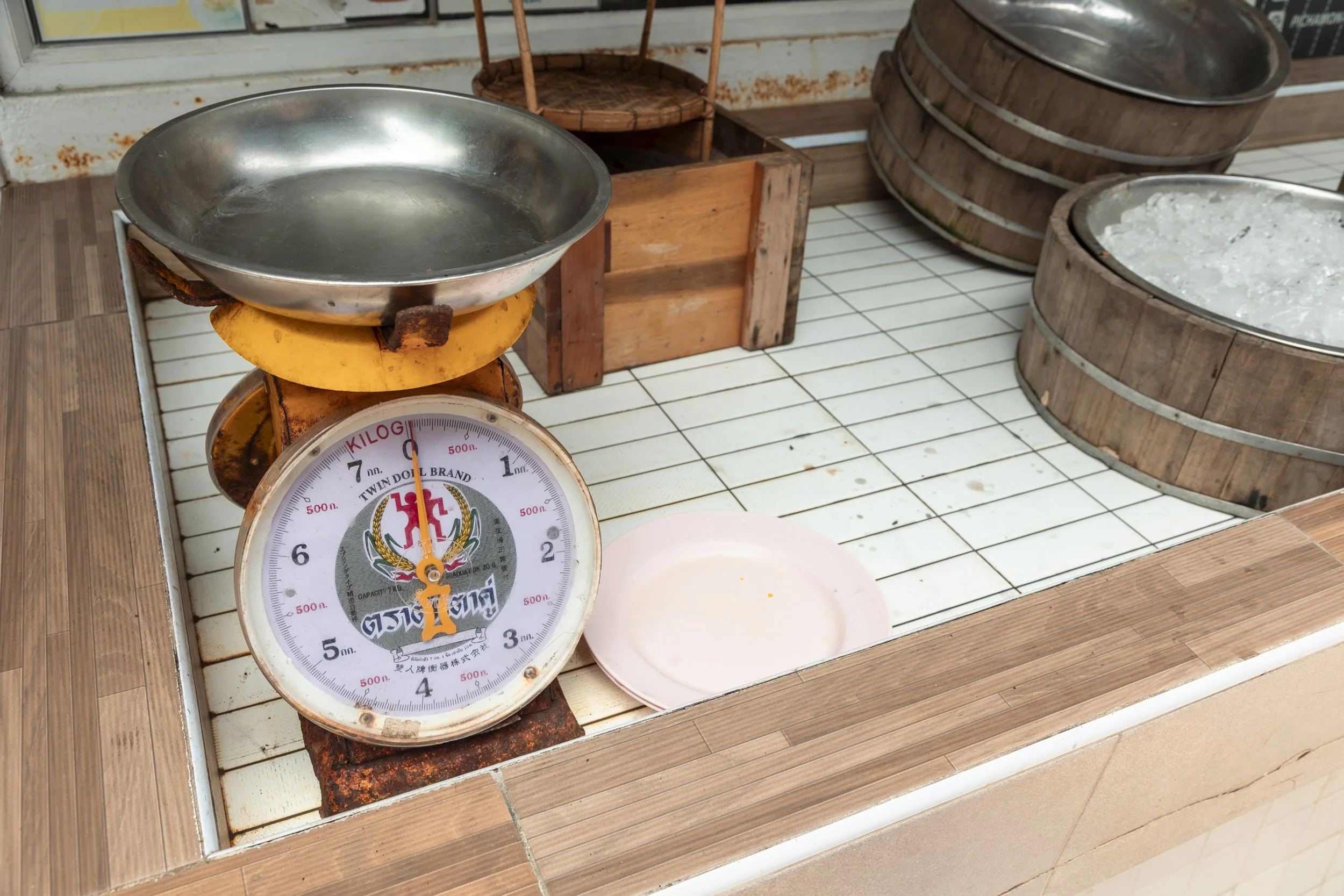An old-fashioned weighing scale on a tiled counter with a metal bowl on top, with another empty plate nearby. Behind the scale are wooden containers, one filled with ice and another with some unidentified substance.