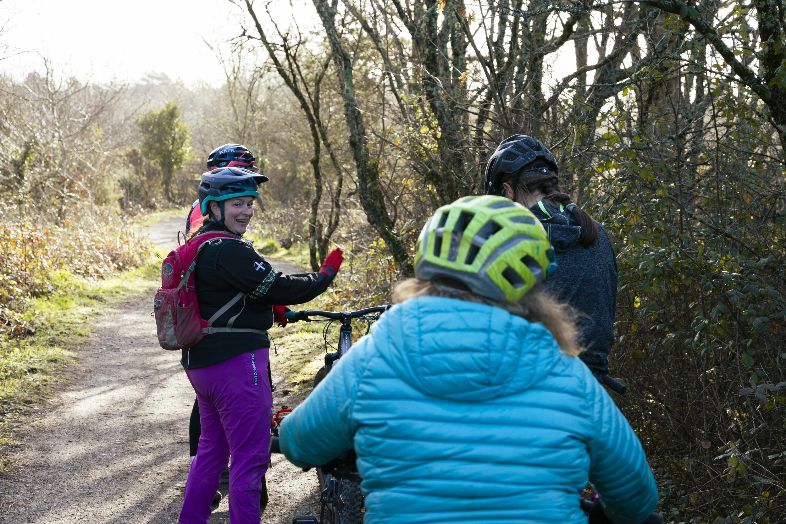 A group of four children in colorful jackets and helmets, with one carrying a pink backpack, are talking and smiling on a forest trail with bare trees and green bushes.