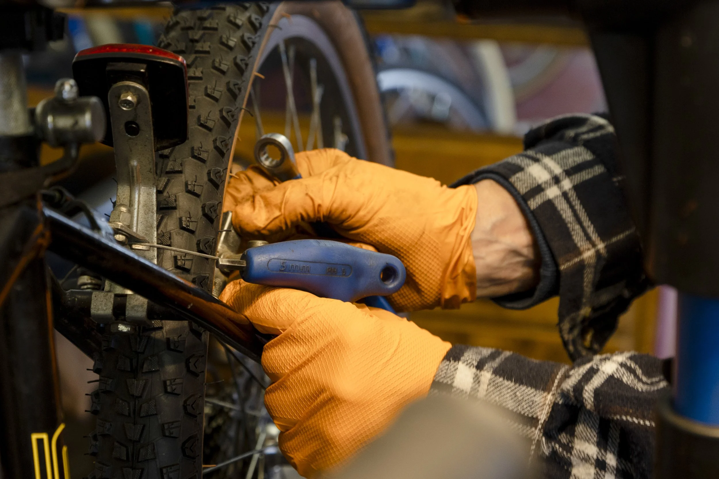 Person working on a bicycle, wearing orange gloves and plaid shirt, tightening a bolt near the rear wheel with a wrench.