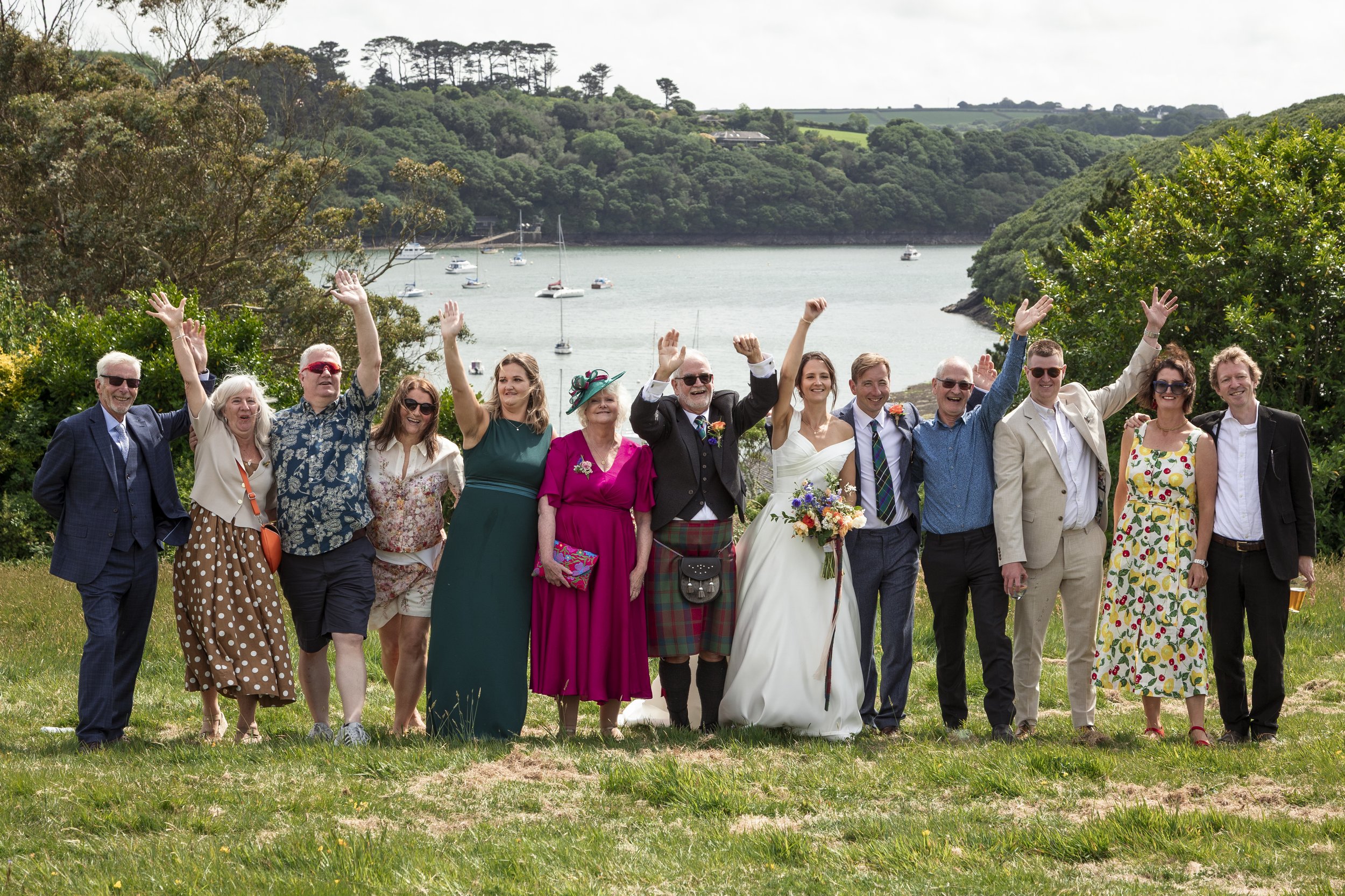 Group of people celebrating outdoors near a body of water with boats, trees, and hills in the background.