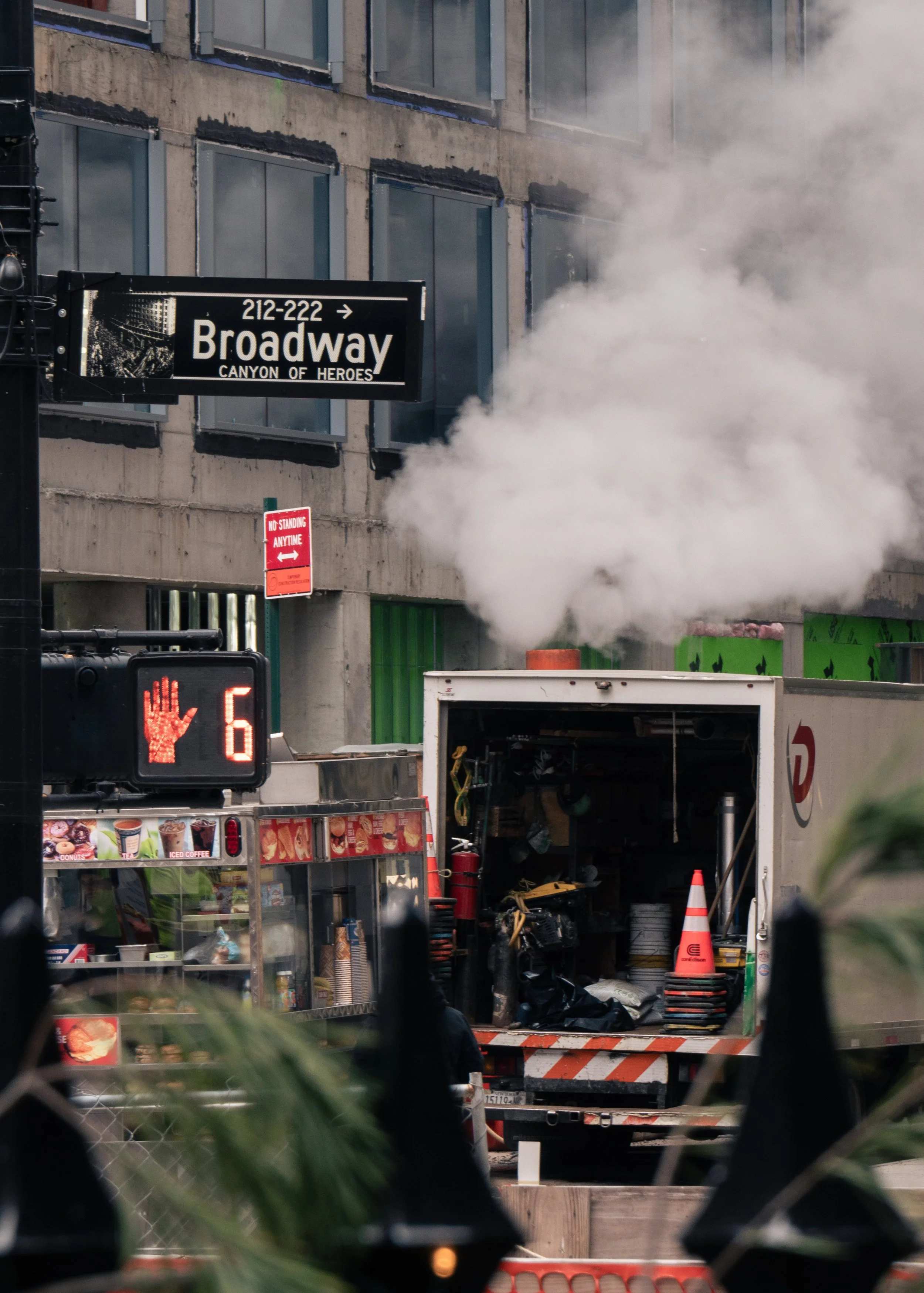 A street scene showing a construction or maintenance vehicle emitting white steam or smoke, with a city street sign for Broadway and a digital traffic signal showing a hand and the number 6. There are buildings and construction barriers visible in th