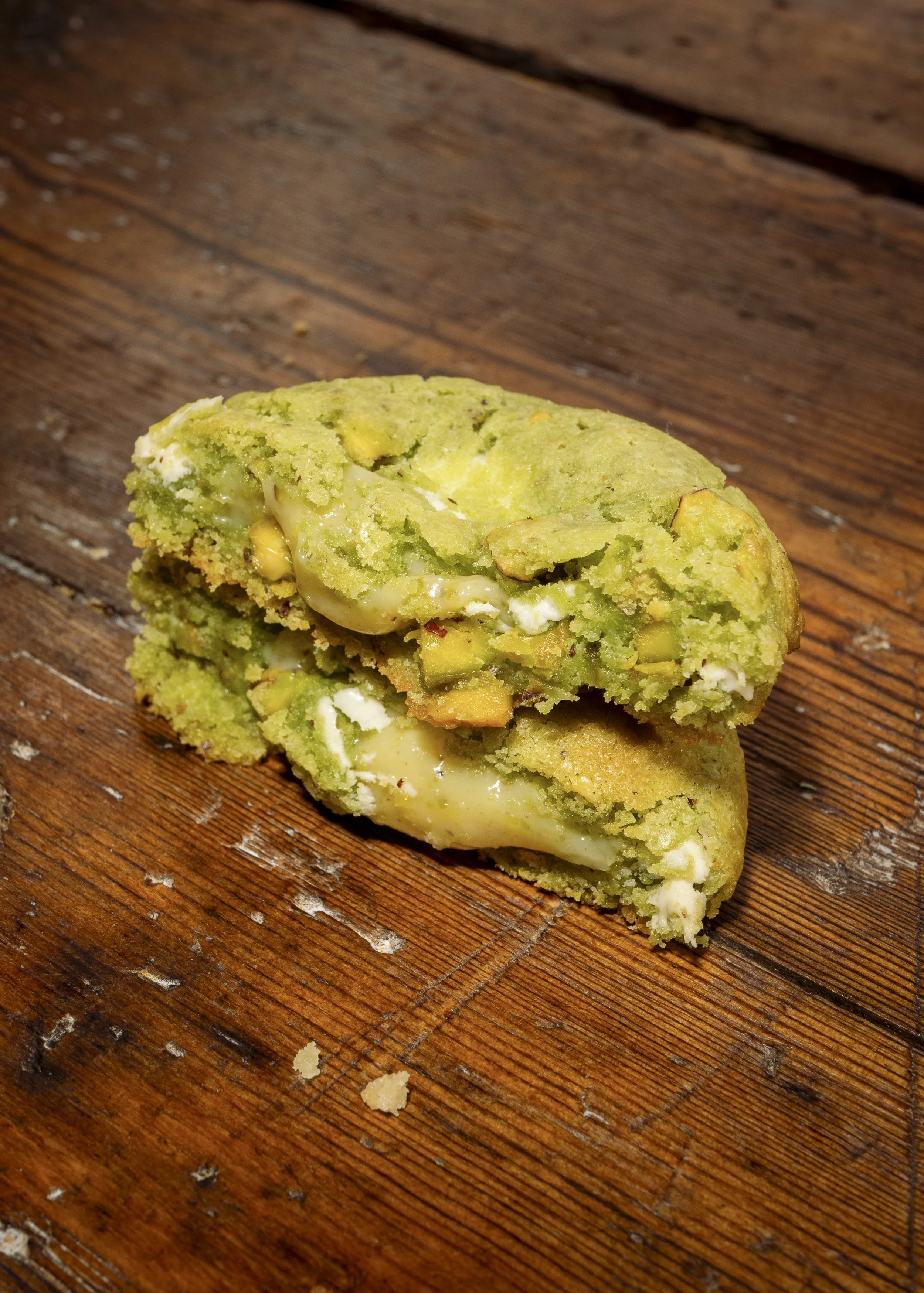 A partially broken green pistachio cookie with creamy filling on a wooden surface.