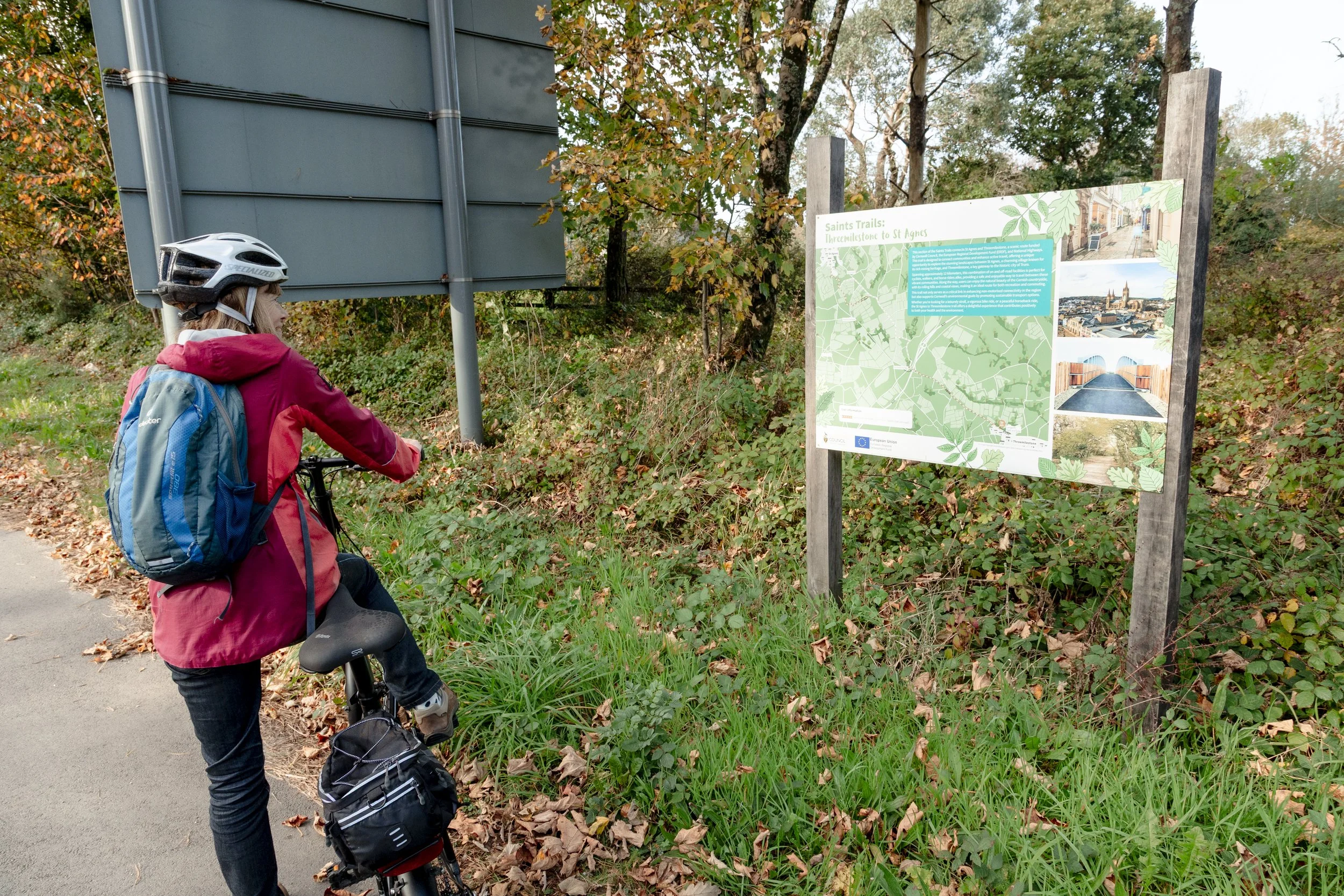 A person with a bicycle, wearing a helmet, red jacket, and a backpack, looking at an informational trail sign on a wooded trail.