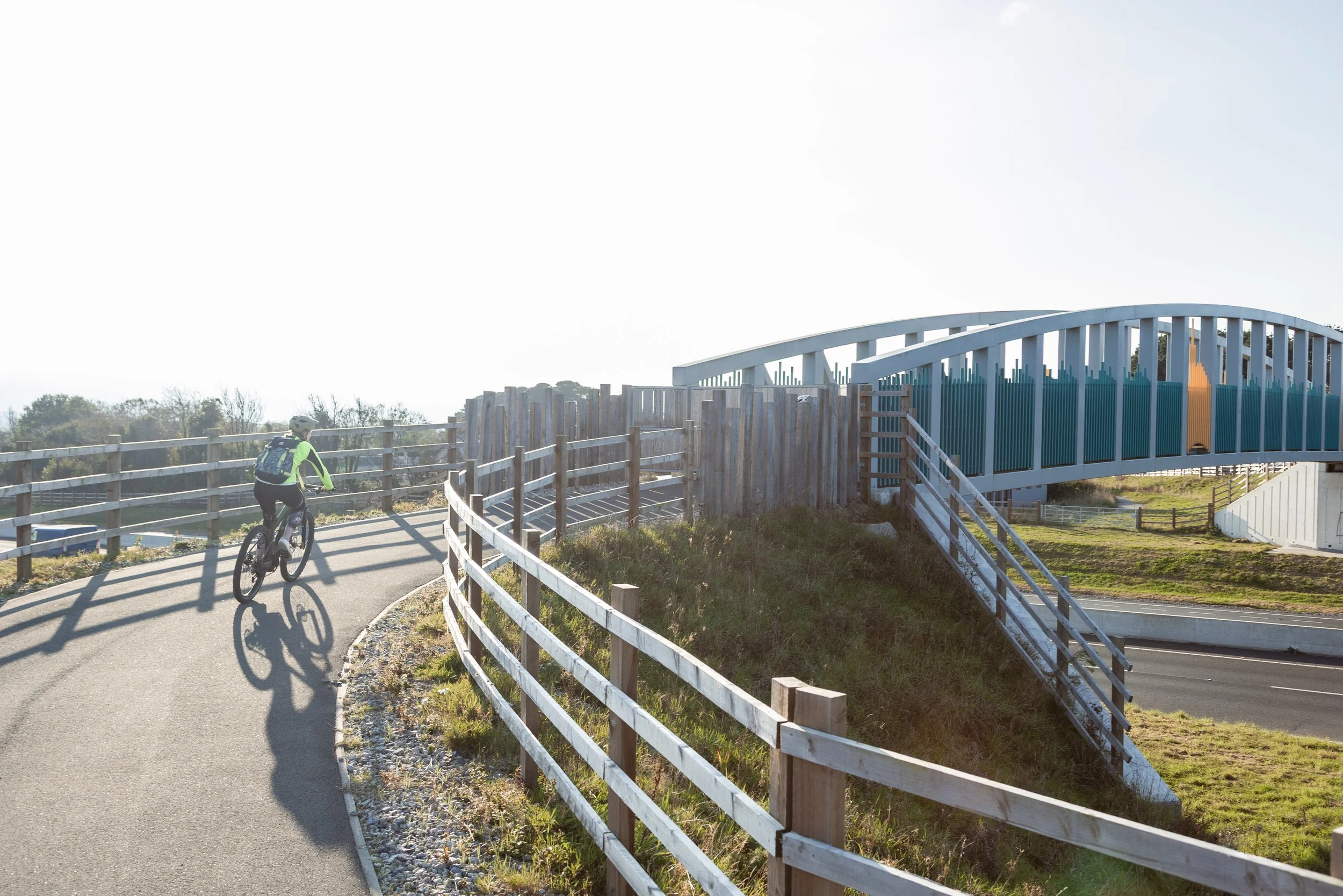 A person riding a bicycle on a paved path next to a wooden fence with a blue and white footbridge overhead, in a park or recreational area on a sunny day.