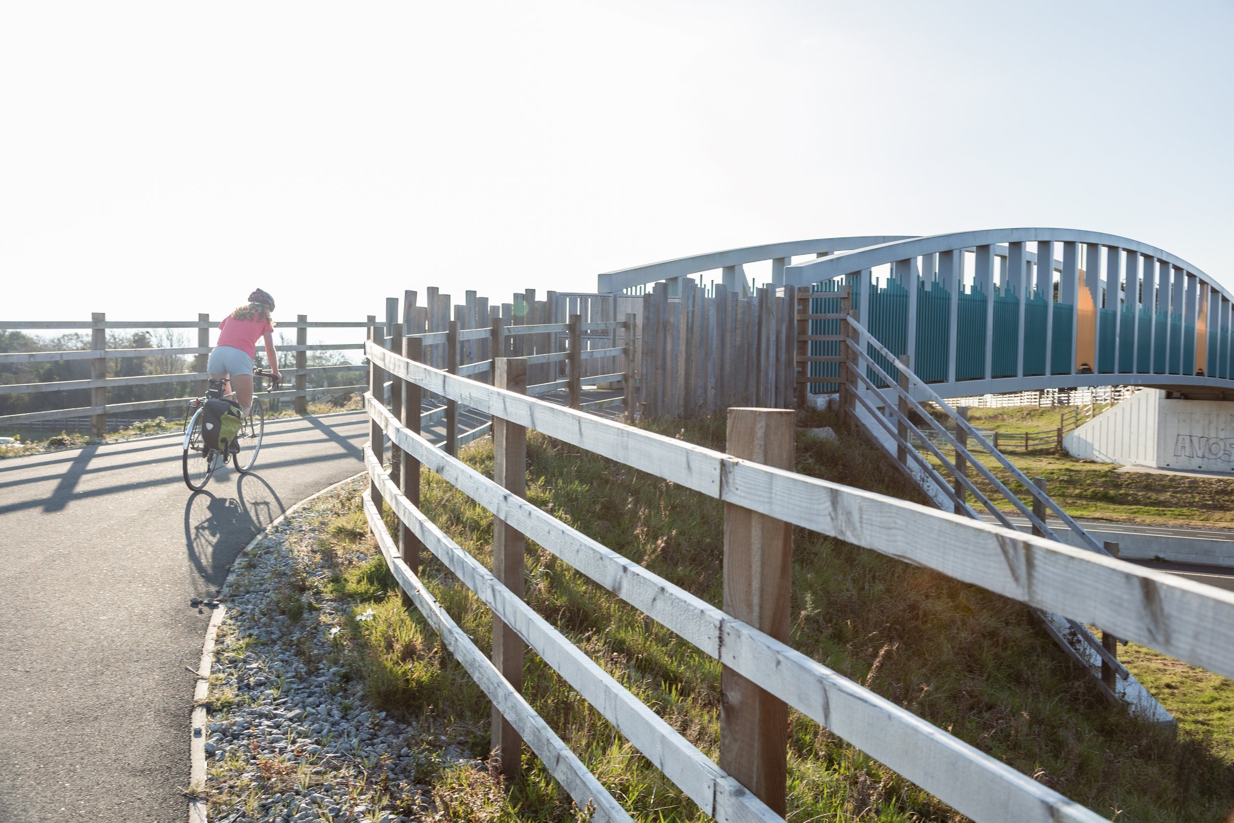 A girl riding a bicycle along a curved paved path, surrounded by wooden fences and an elevated bridge structure in a sunny outdoor setting.