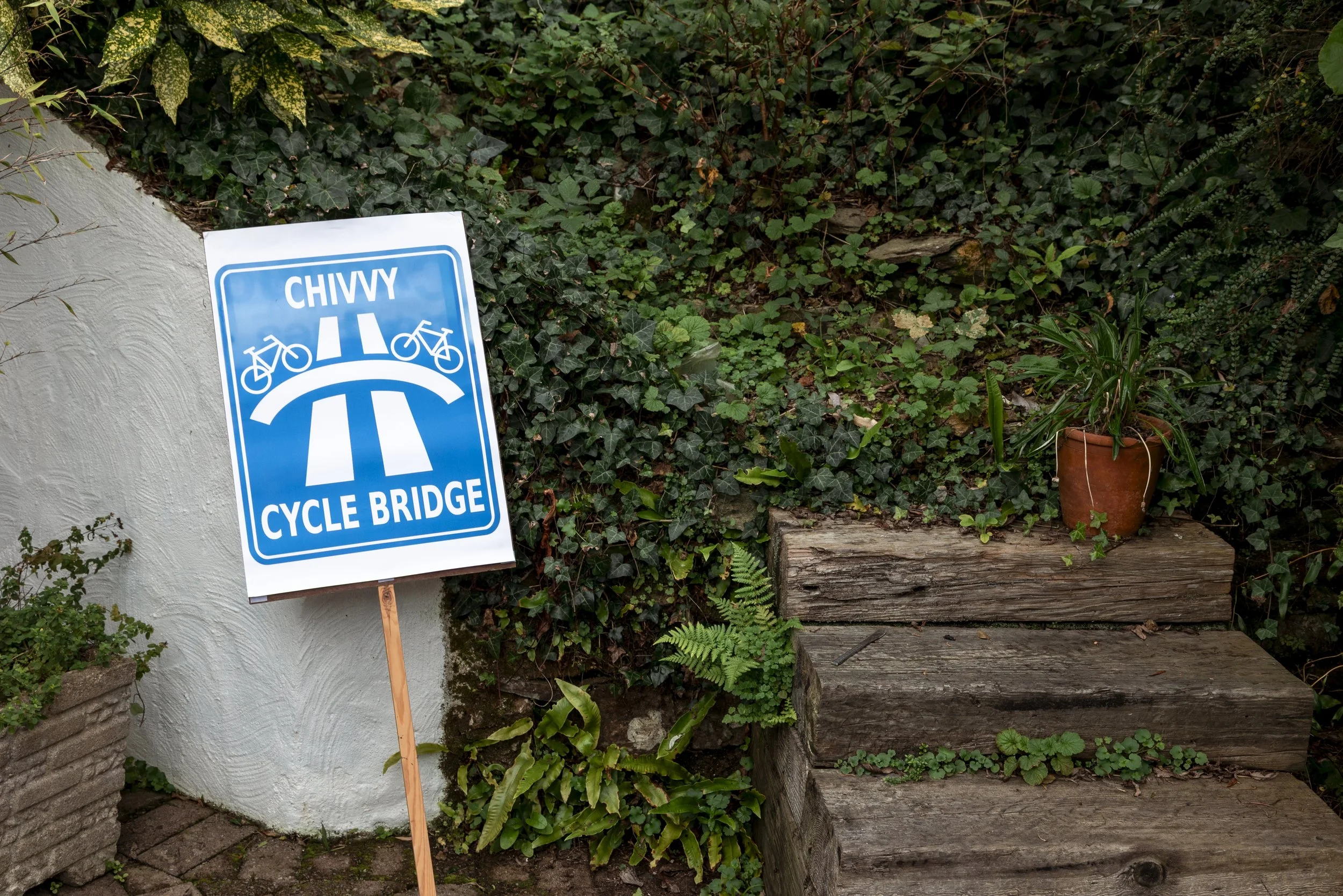 A sign indicating a cycle bridge with the word 'Chivy' and symbols of bicycles on a bridge. The sign is leaning against a wall next to wooden steps and a pot with a green plant.