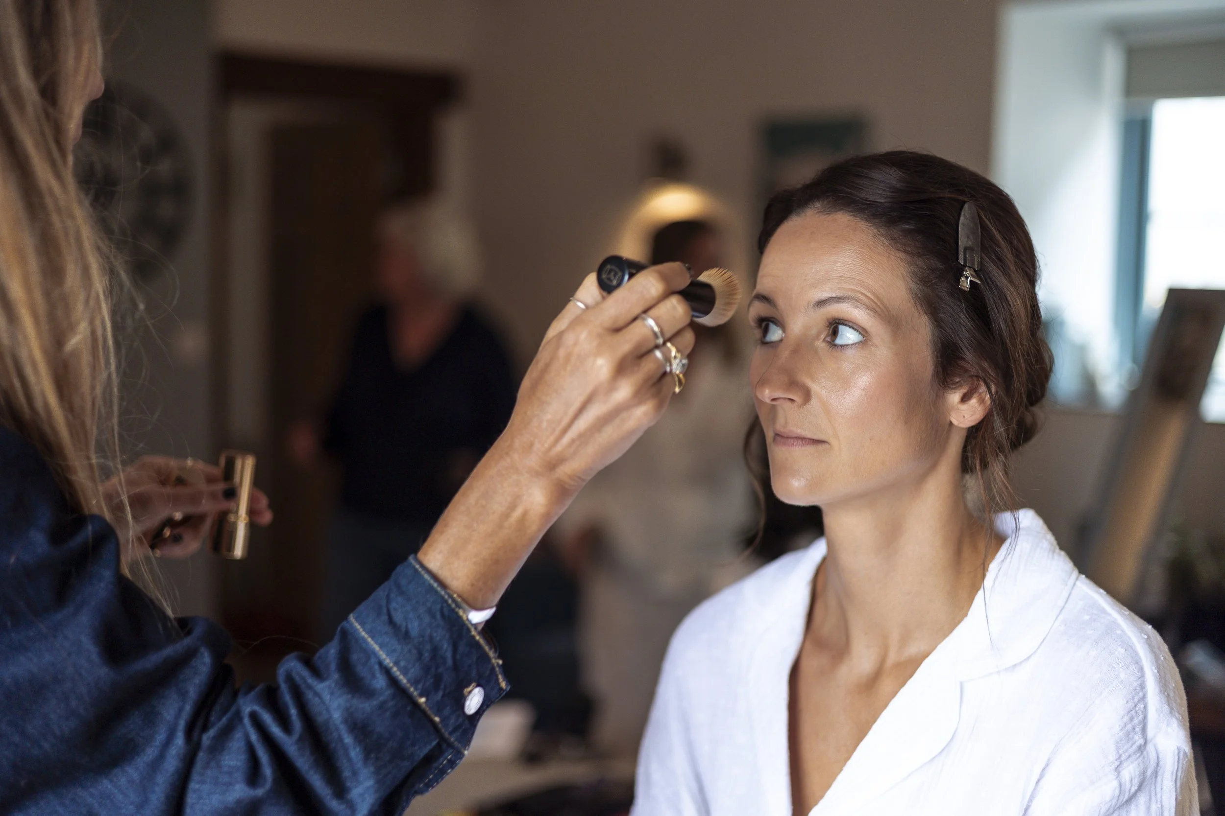 A woman sitting for makeup application by a makeup artist in a room, with two women in the background.