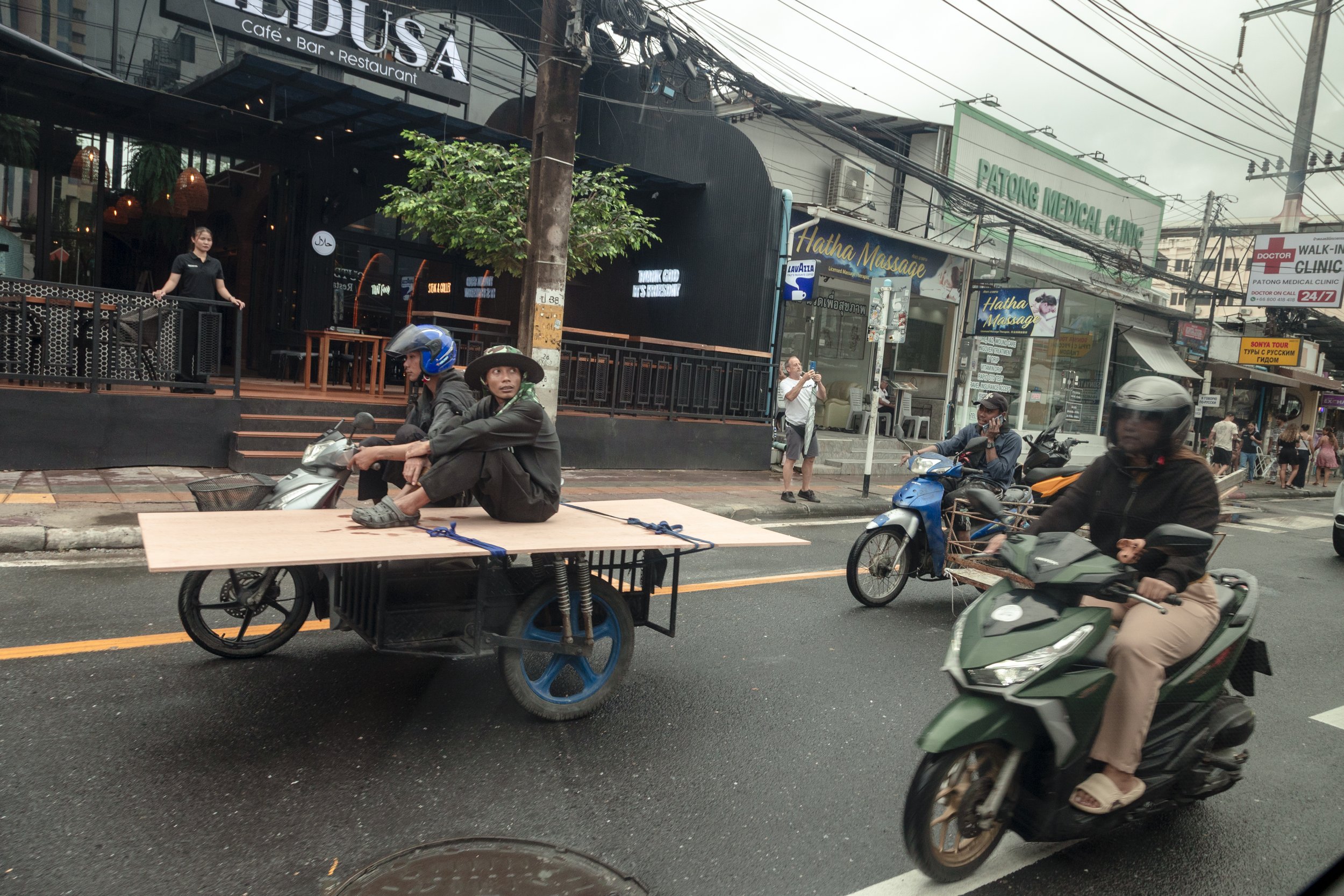 Street scene in an urban area with motorcycles and pedestrians, storefronts on the sidewalk including a restaurant called 'ENDEUS' and a medical clinic sign, with electrical wires overhead.