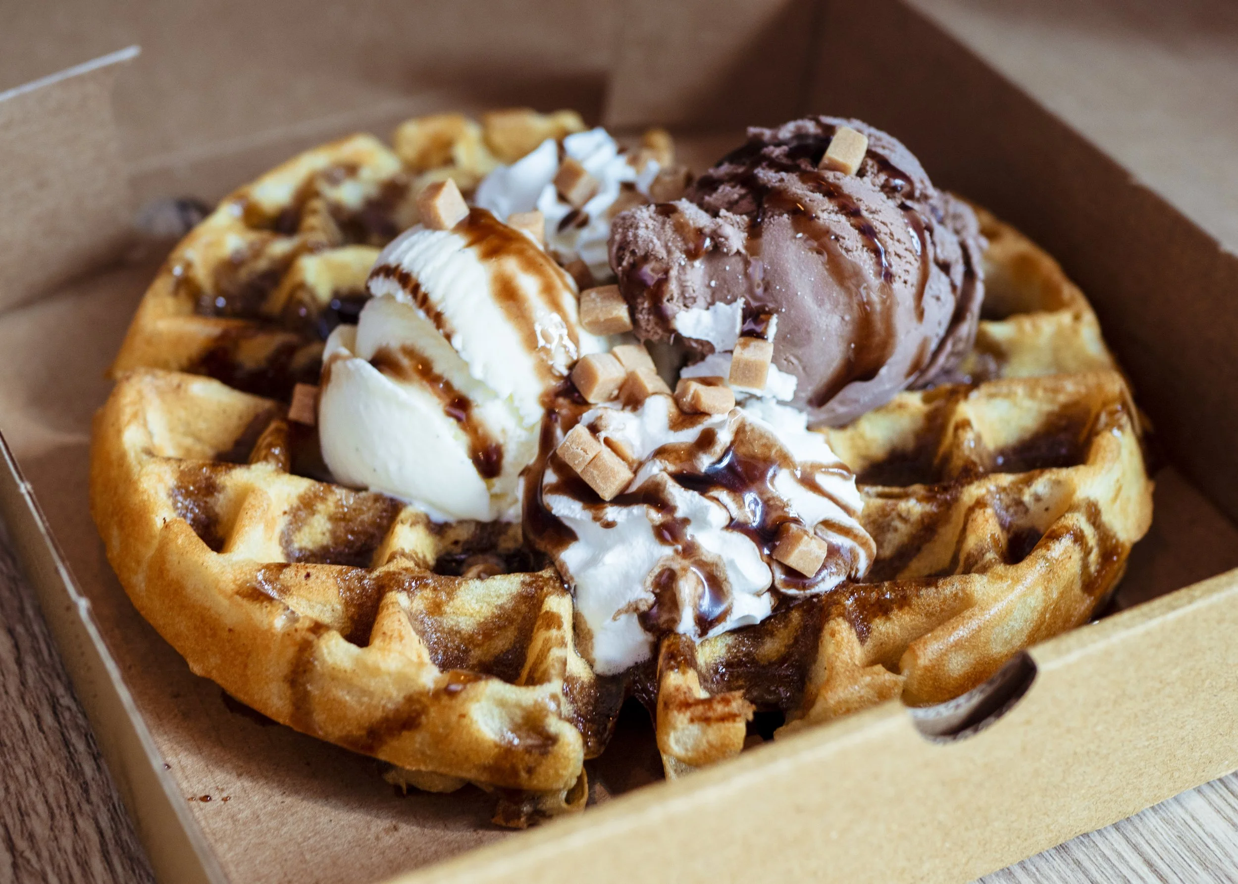 Waffle topped with vanilla and chocolate ice cream, whipped cream, caramel sauce, and caramel cubes in a brown takeout box.