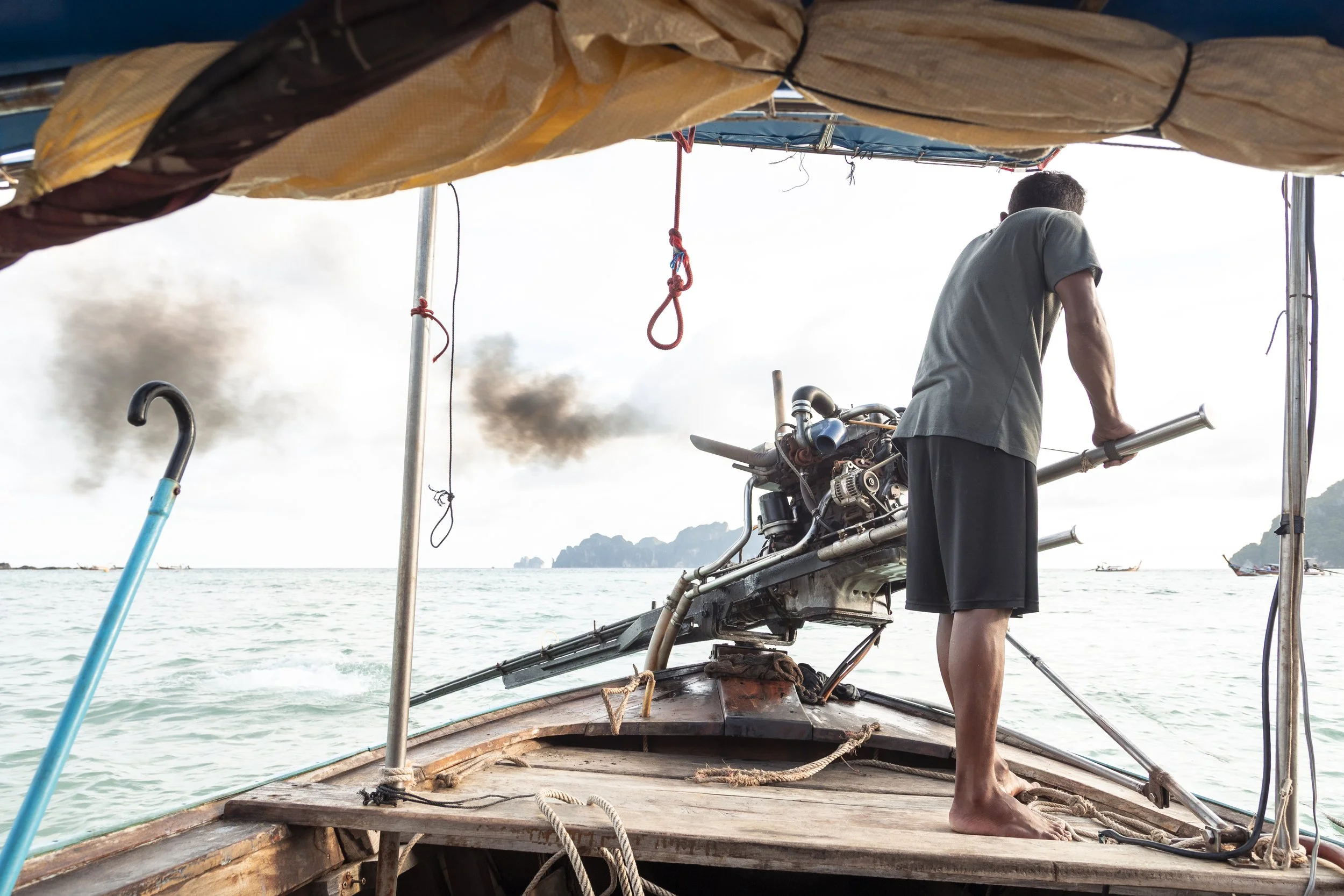 A man standing barefoot on the deck of a boat, holding onto a metal pole, with a damaged outboard motor, black smoke emitting from near the water, and distant islands on the horizon.