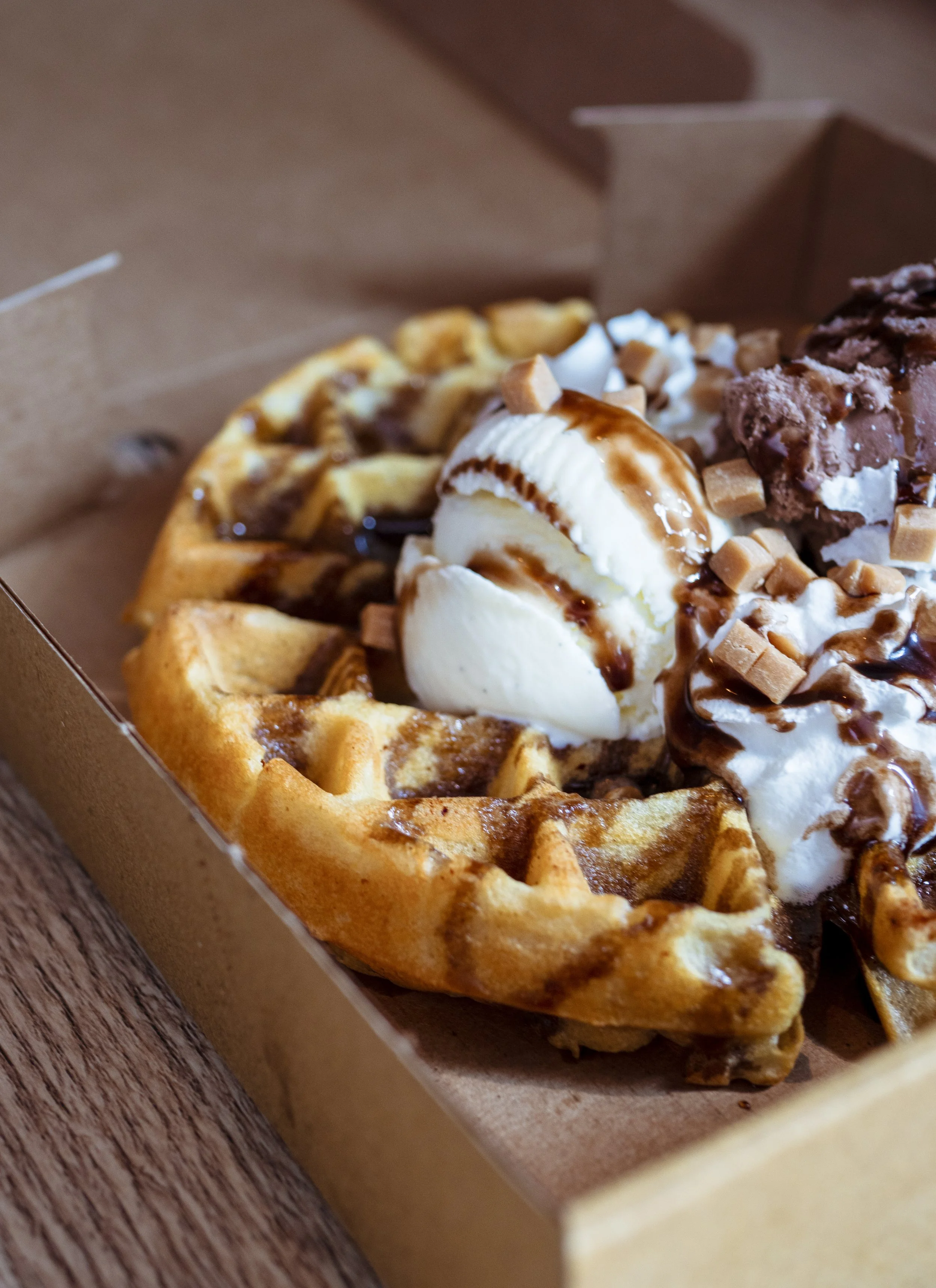 A dessert pizza with waffle crust, topped with vanilla ice cream, caramel sauce, whipped cream, and caramel candies, in a takeout box on a wooden surface.