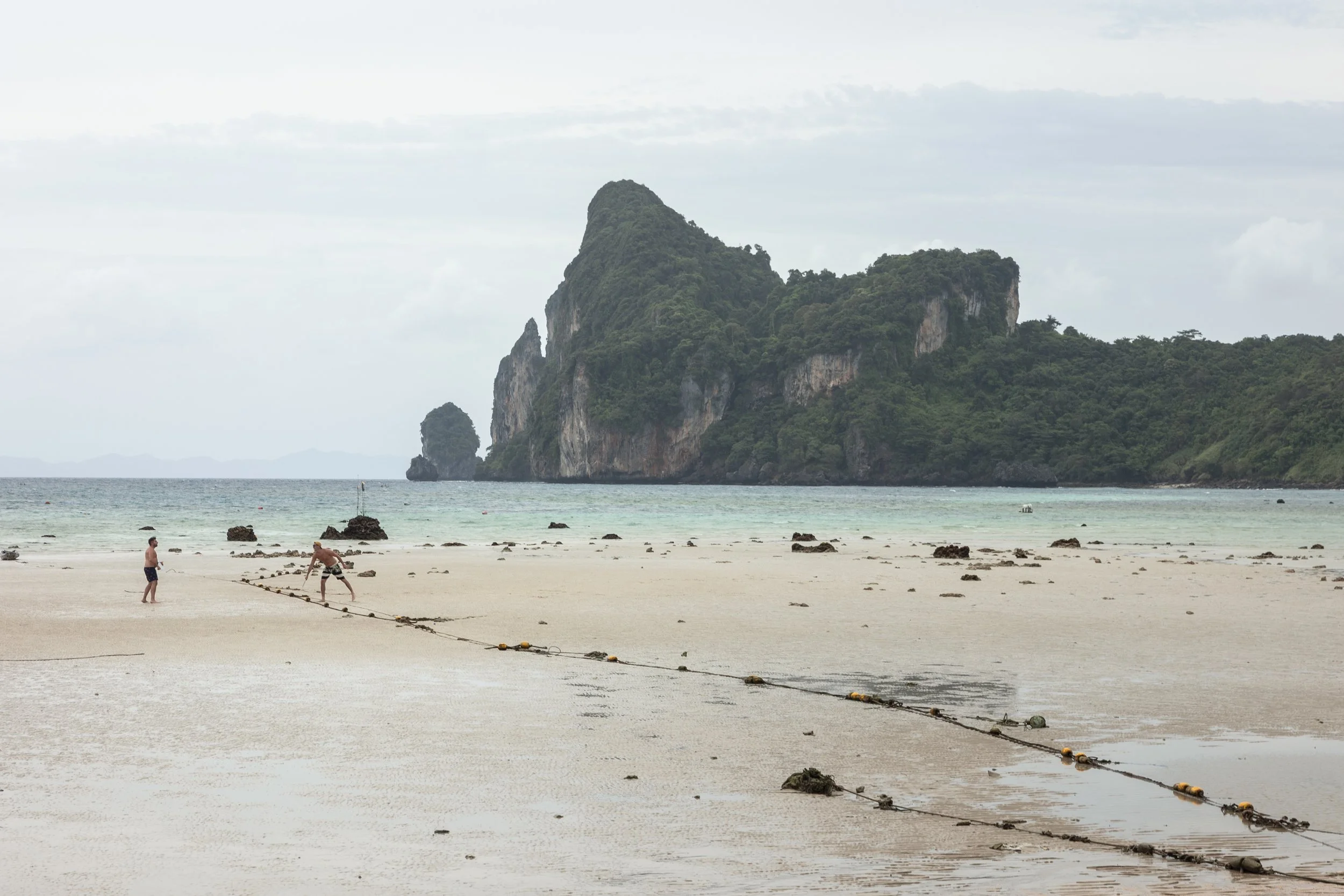 Two men on a sandy beach working with a rope as they prepare for a water activity, with lush green islands in the background and a cloudy sky overhead.