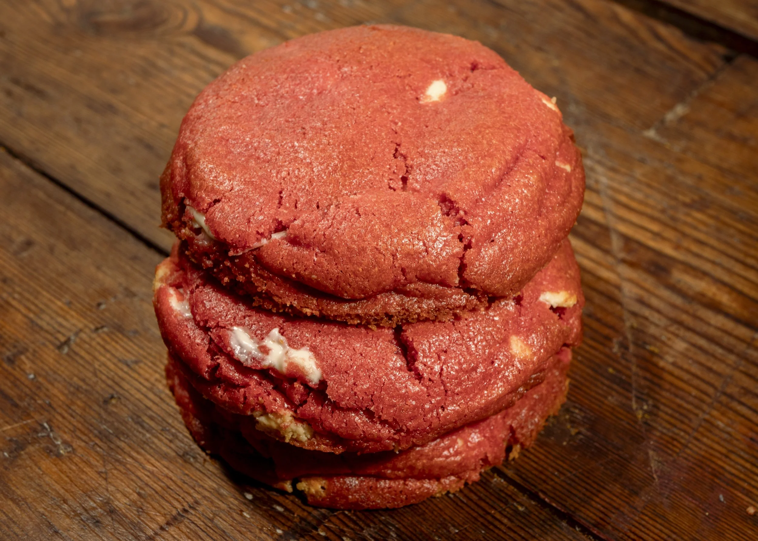 Three red cookies stacked on a wooden surface, with white chocolate chunks visible in some.