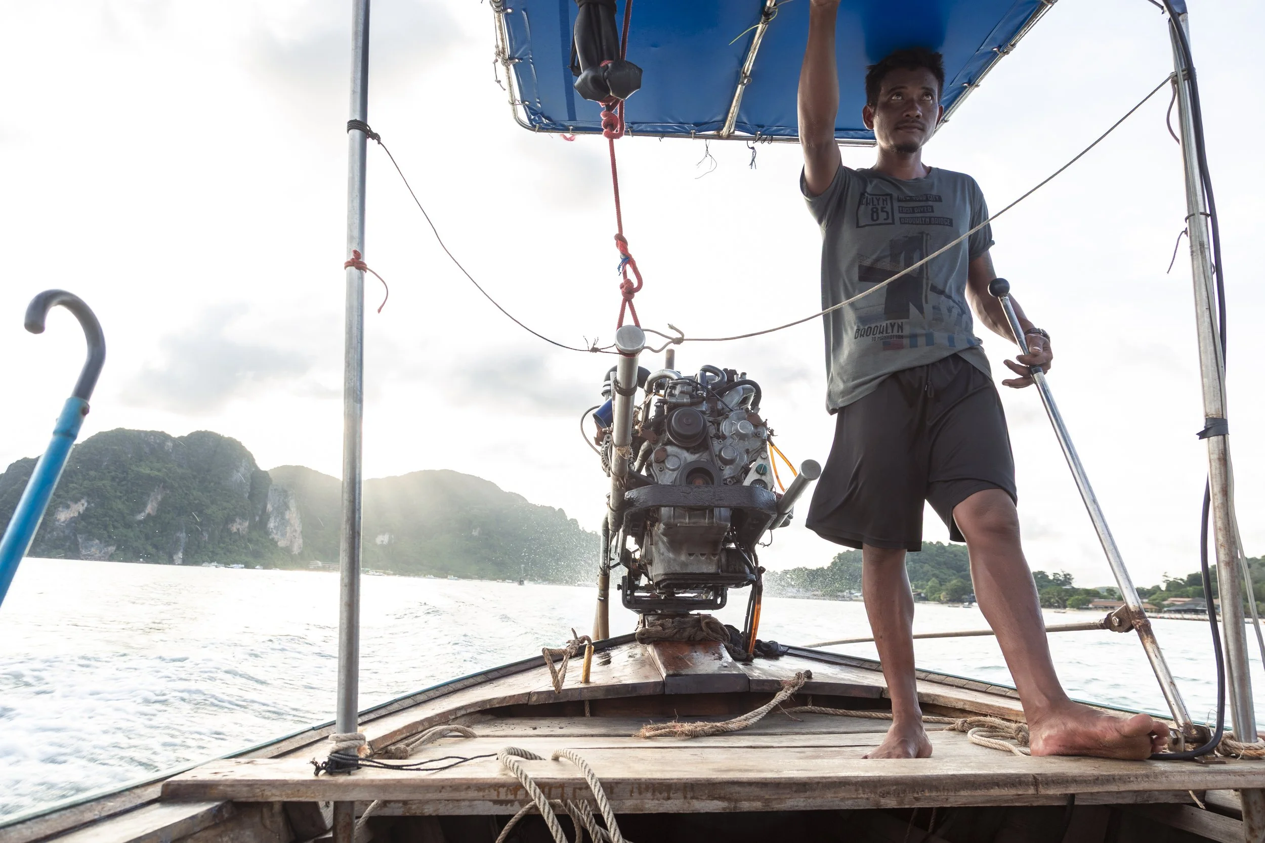 A man standing barefoot on the front of a boat operating an engine with a scenic view of water and lush green islands in the background during daylight.