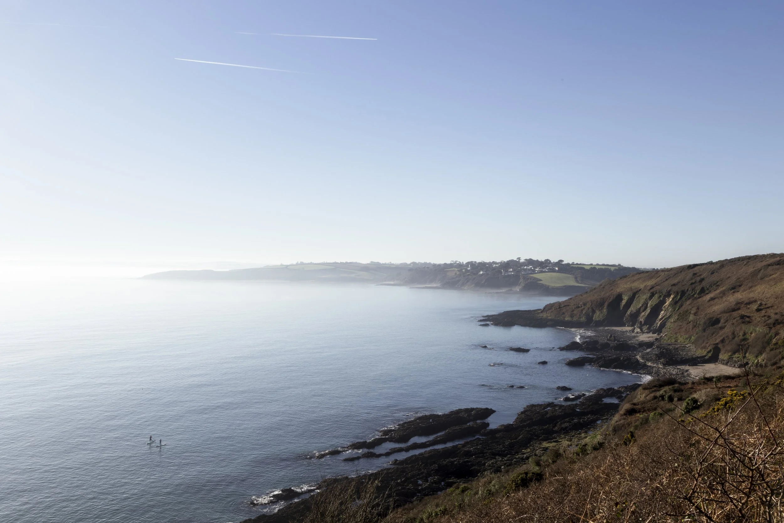 Scenic view of a distant shoreline with cliffs, calm ocean water, and two paddleboarders. Clear sky with a contrail visible.