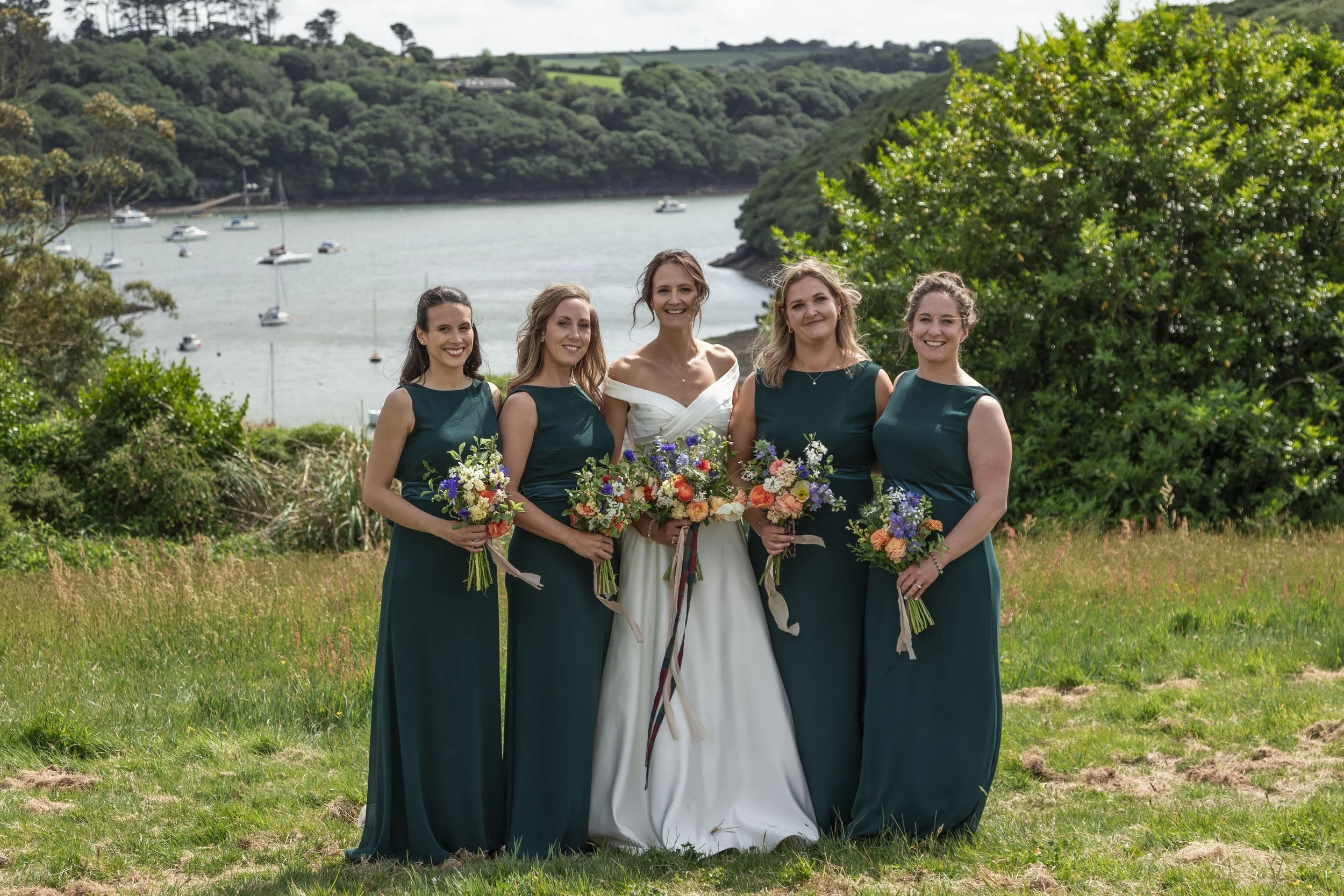 Group of five women in dresses, standing outdoors on grass with water and trees in the background. One woman in a white wedding dress, four women in dark teal dresses, holding bouquets of flowers.