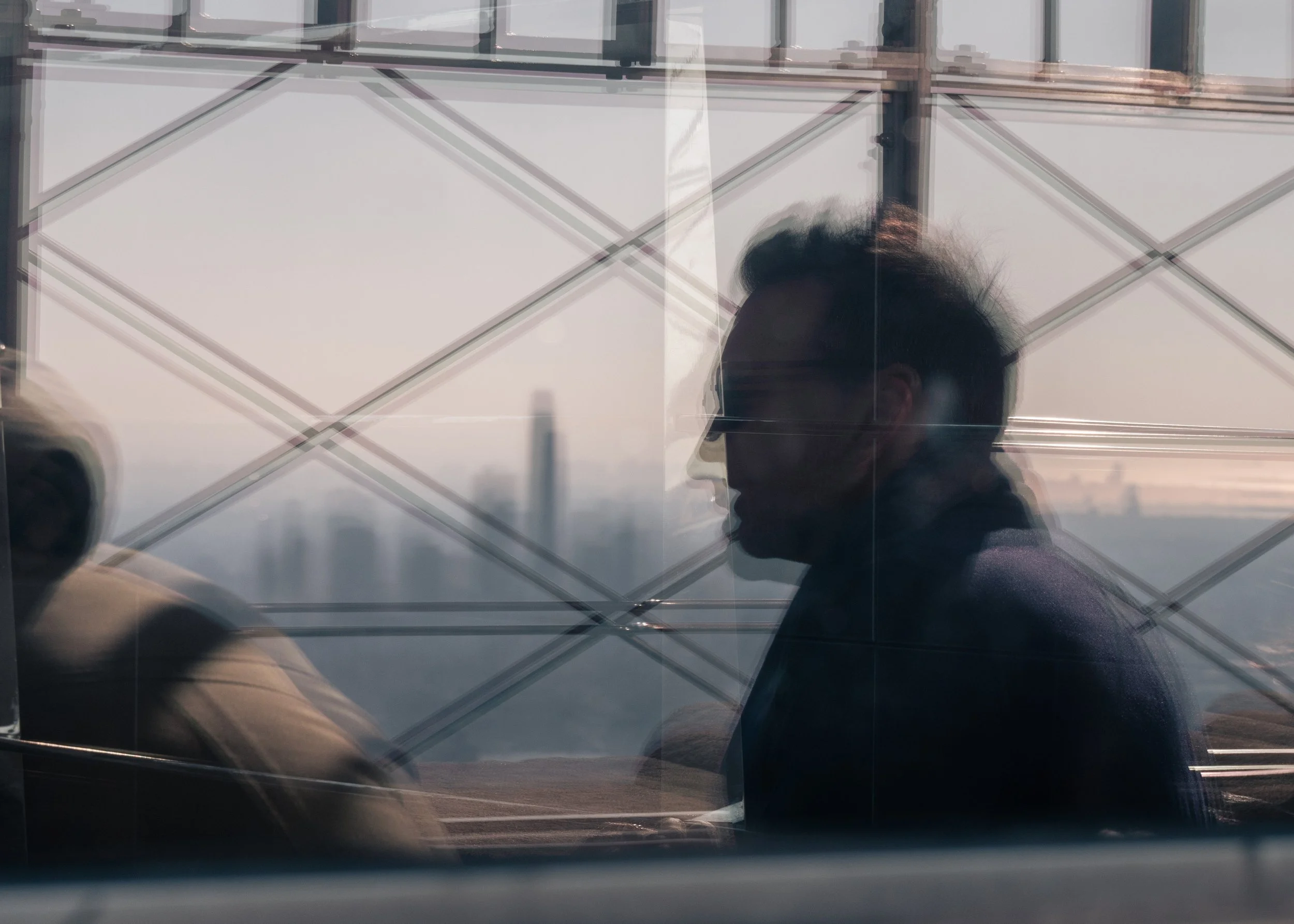 A silhouette profile of a man wearing glasses, sitting inside a building with a reflective glass window, through which an urban cityscape is visible in the background.