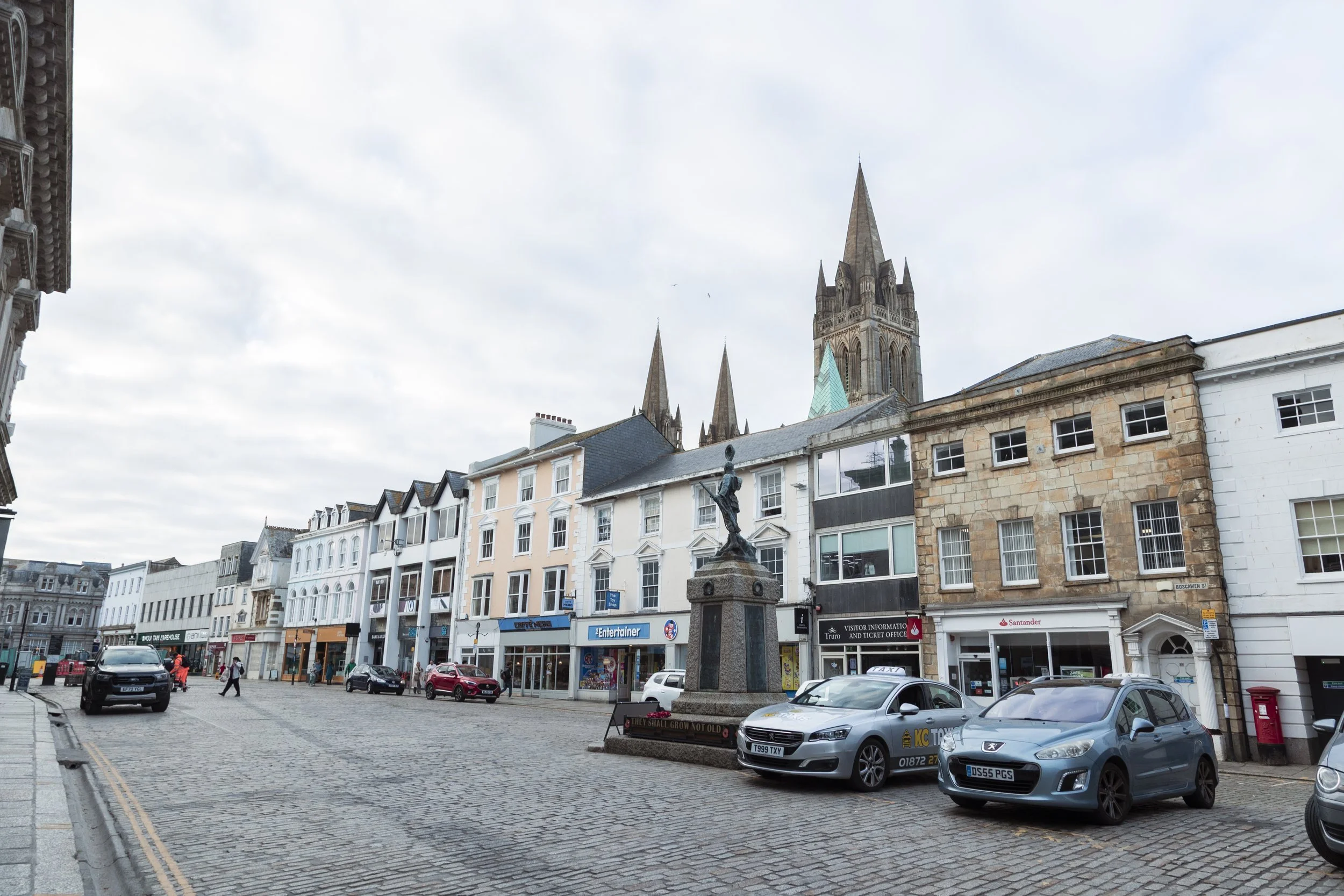 A cobblestone street in a city center with parked cars, shopfronts, and a statue of a woman. In the background are historic church spires.