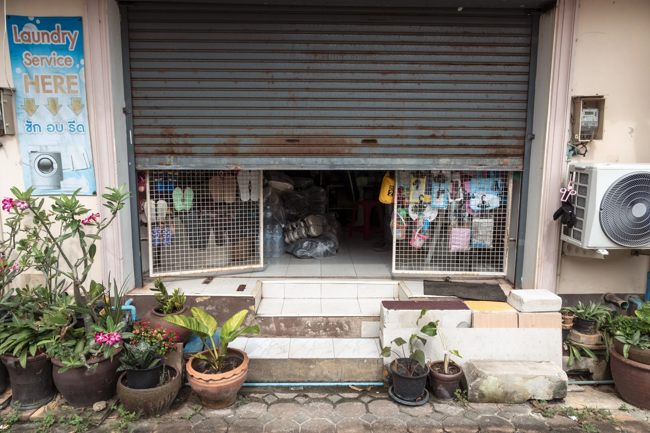 A small shop or laundry with a partially open rusty metal shutter. Potted plants and flowers are placed in front of the shop. Inside, various items are visible, including water bottles and small household goods. A sign on the left indicates laundry s