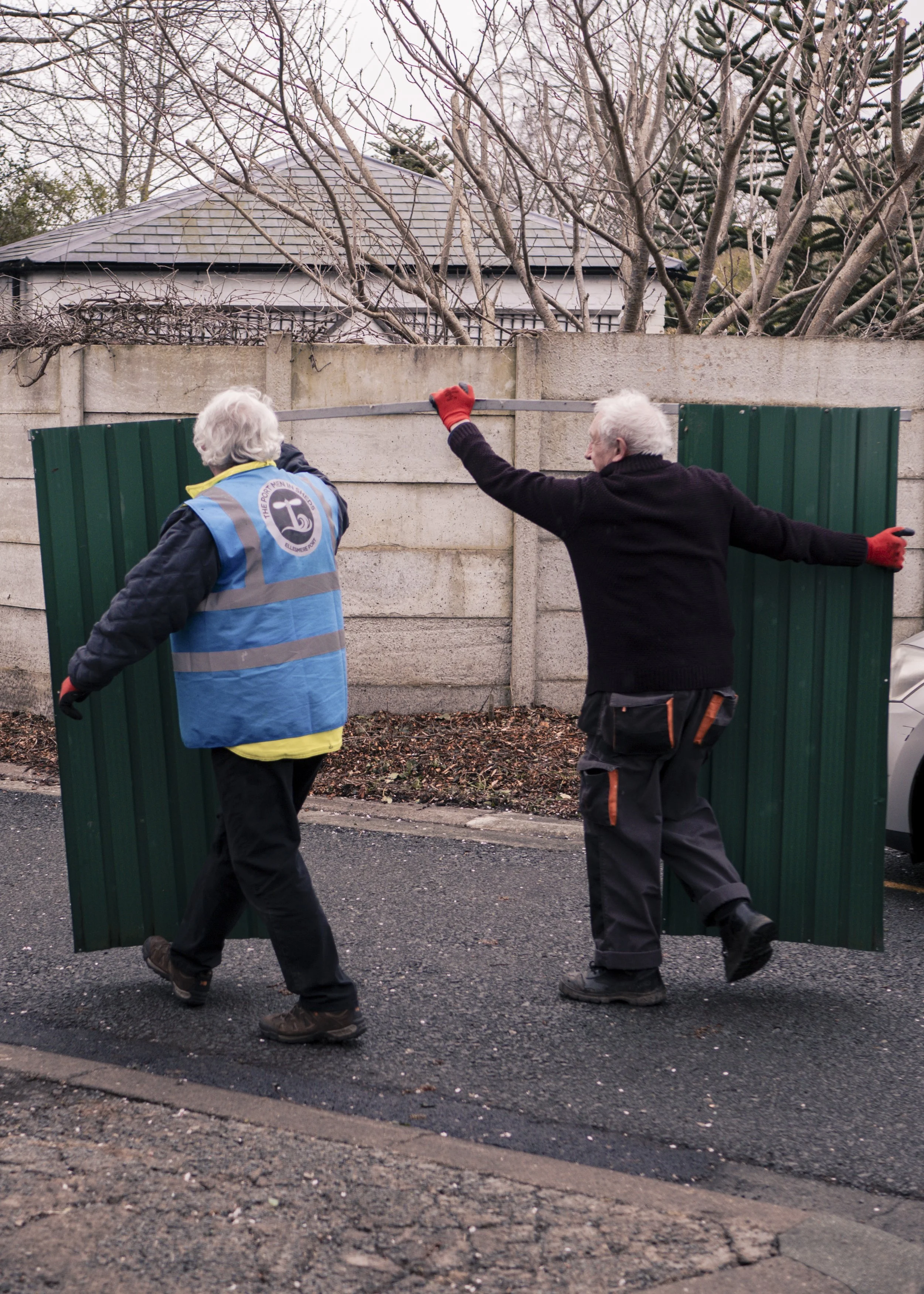Two elderly men are installing a green metal fence panel along a concrete wall on a street. One man is wearing a high visibility vest, and both are wearing gloves.