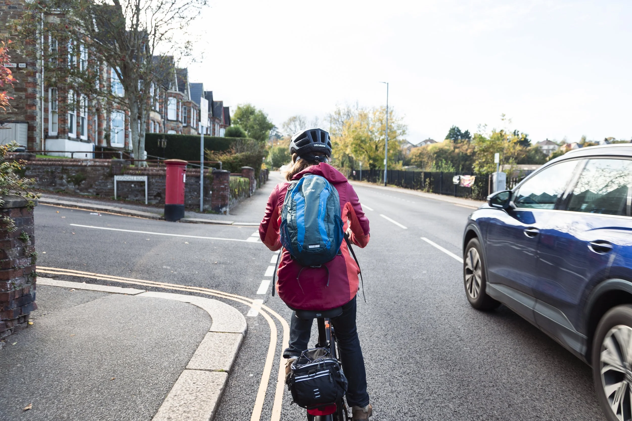 Person riding a bike on a city street, wearing a black helmet and a red jacket, with a blue backpack, passing parked cars and residential buildings in the background.
