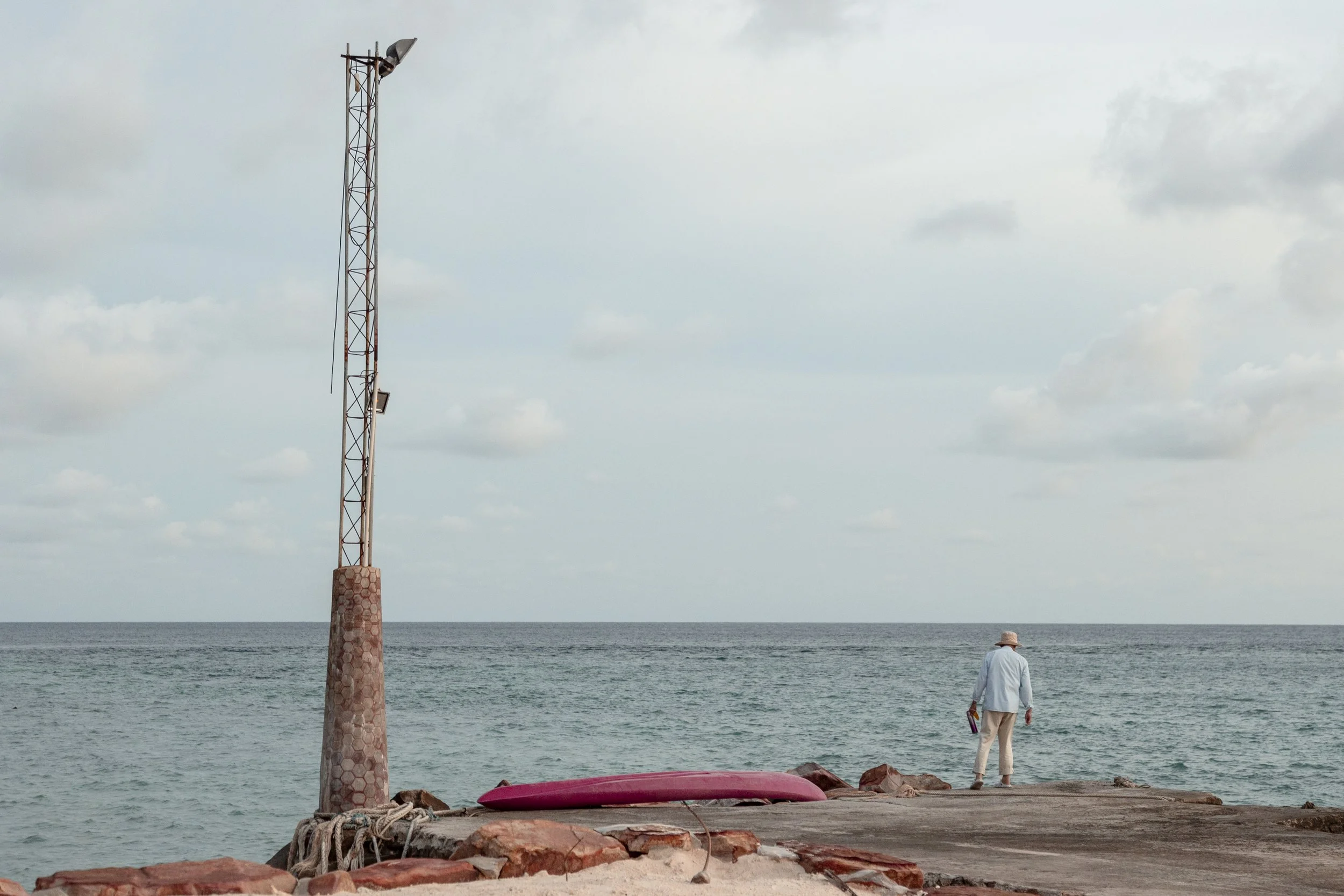 A person stands on a rocky pier near the ocean, facing the water. A pink kayak is lying on the ground, and there is a tall, rusty pole with a light or camera mounted on top. The sky is partly cloudy.