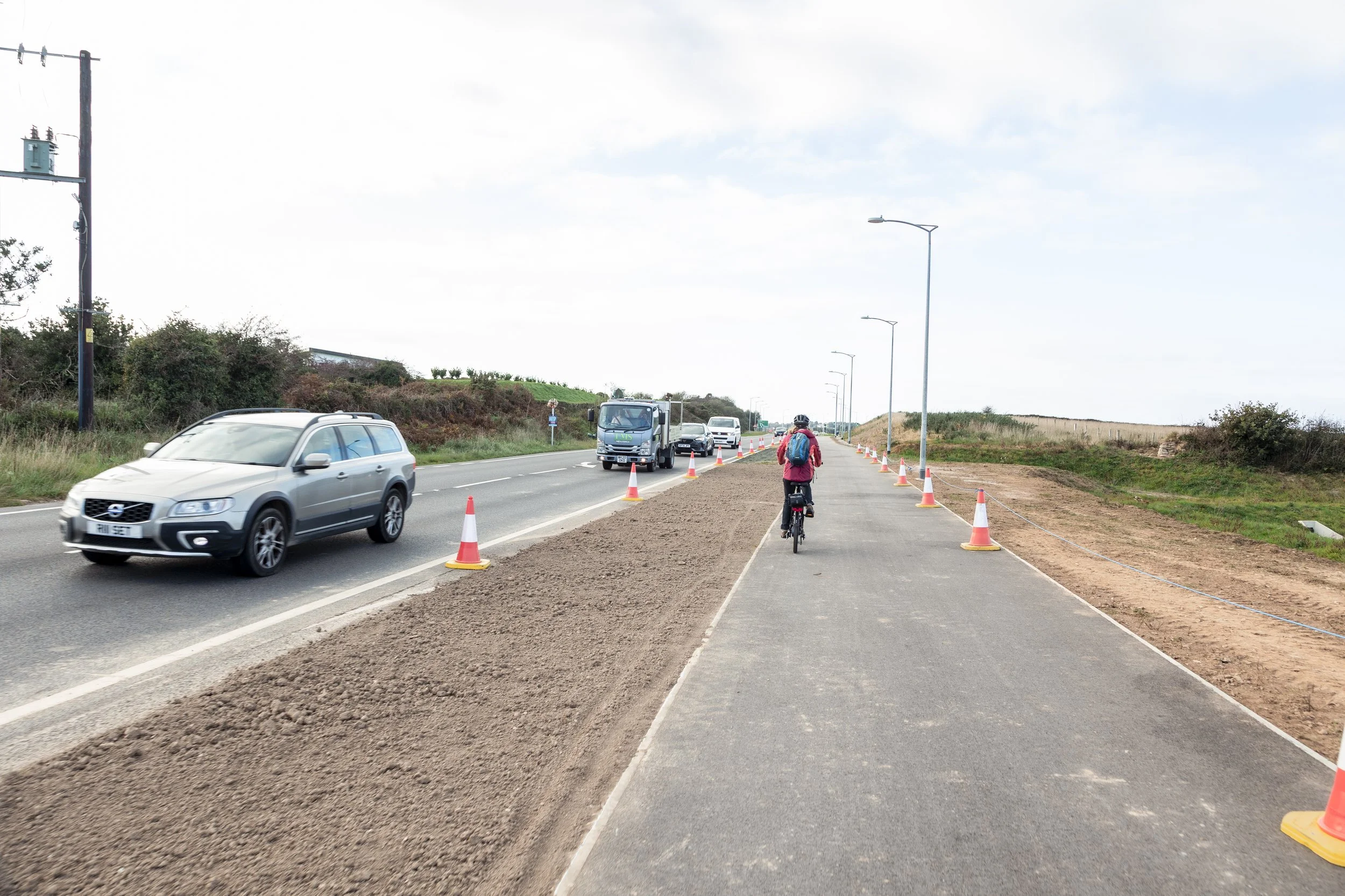 A person riding a bicycle on a paved bike lane next to a highway with cars and trucks. Cones mark the edge of the bike lane, and there are street lights along the road on a partly cloudy day.