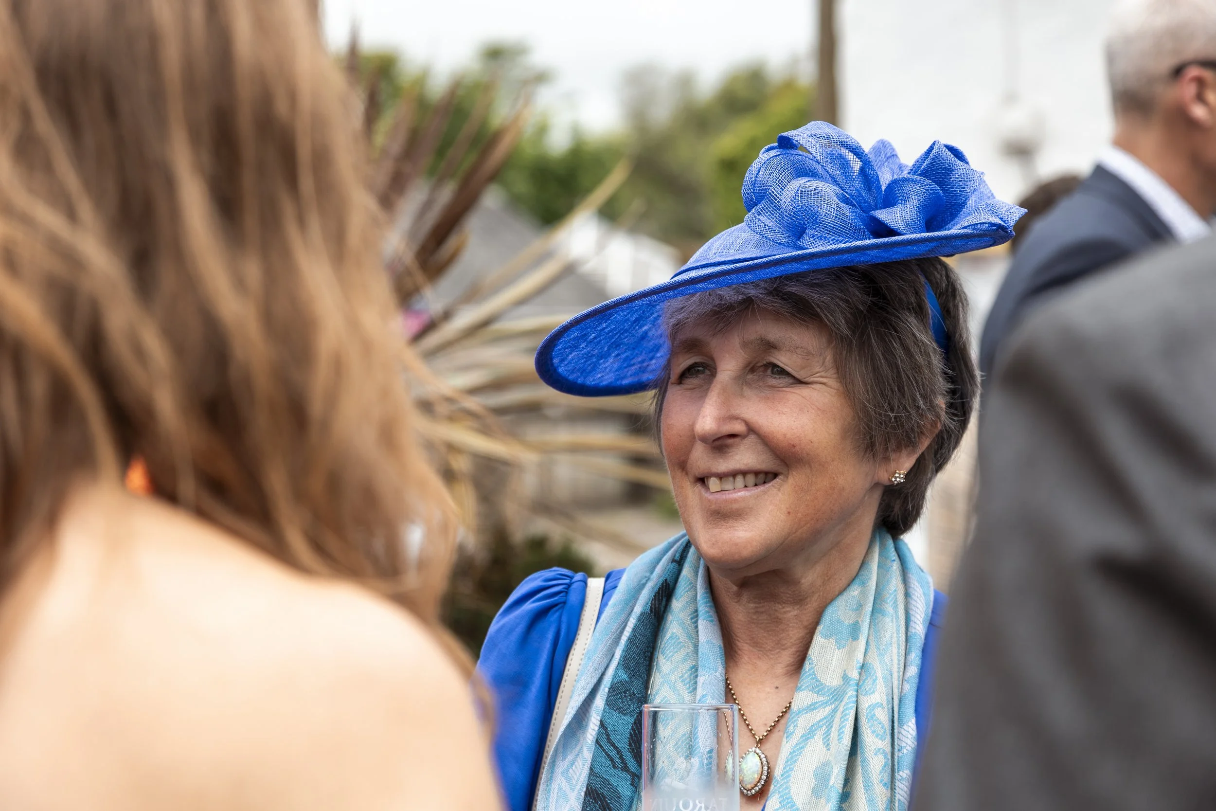 A woman in a bright blue dress and matching hat with netting, smiling and holding a glass at an outdoor event surrounded by people.