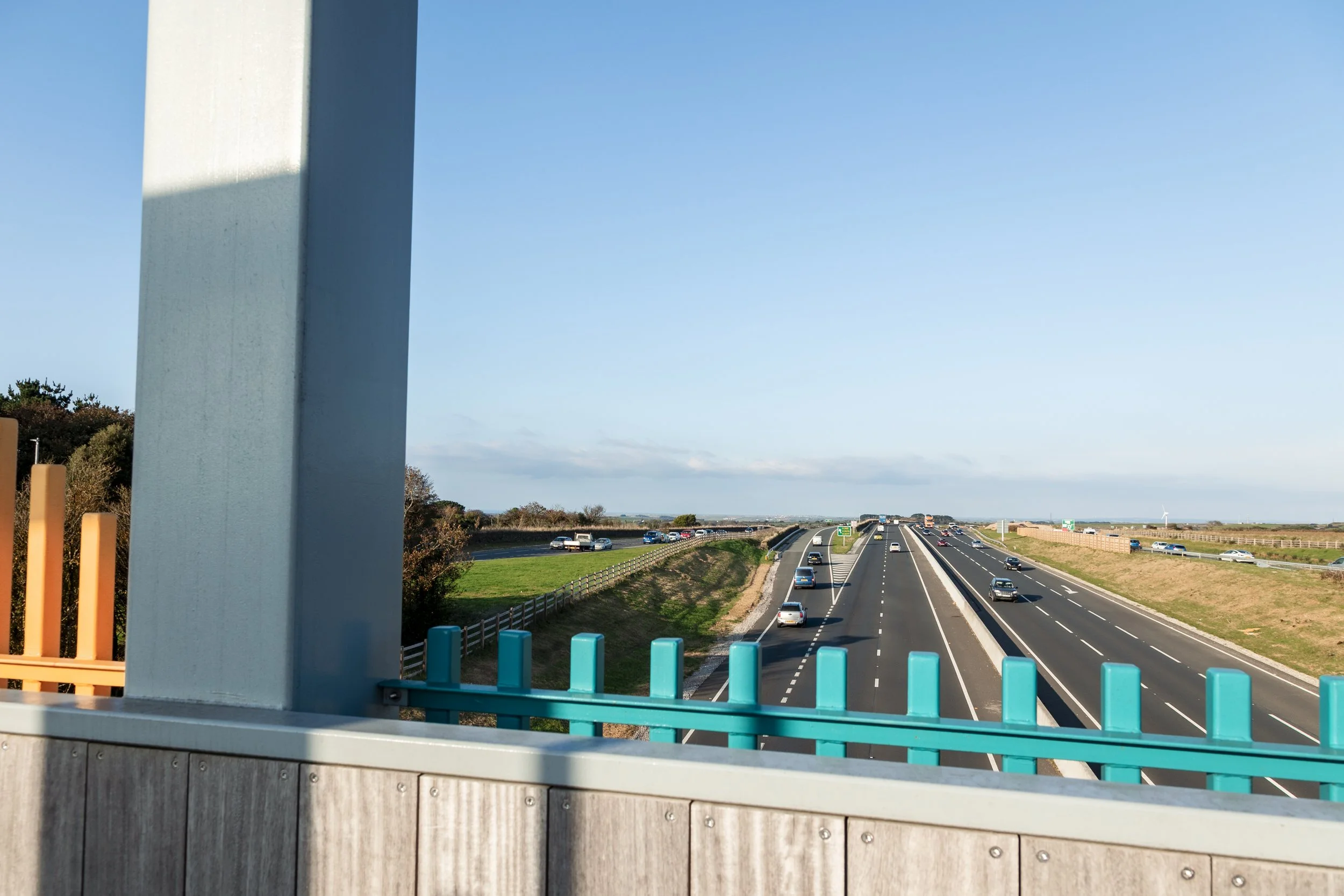 View of a highway with multiple lanes, cars traveling in both directions, and blue sky with few clouds. A sidewalk with a colorful fence and a large vertical support structure are in the foreground.