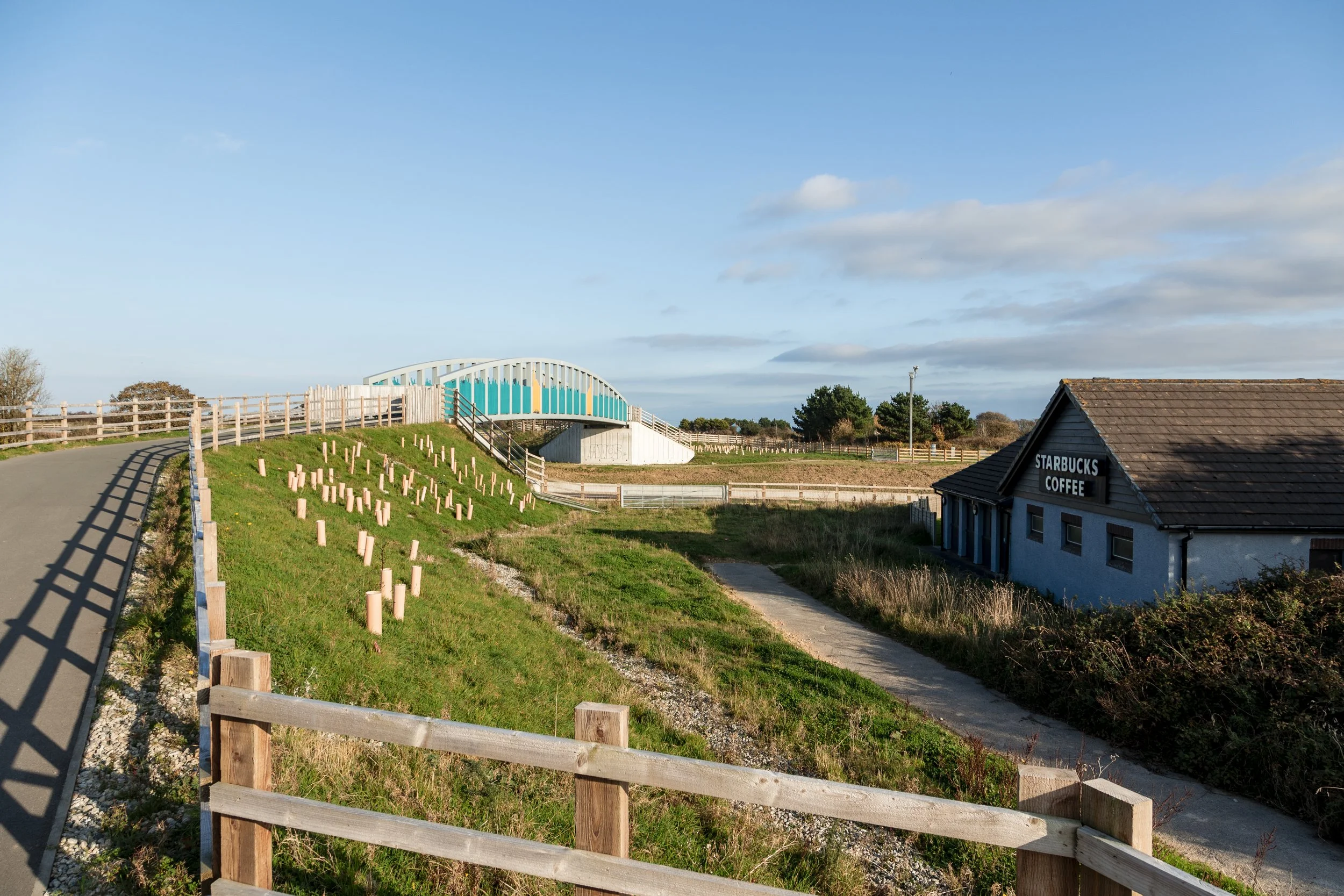 A scenic view of a small road leading to a modern bridge with a blue and white design, a grassy hill with wooden posts, a small concrete path, and a Starbucks Coffee shop with a dark roof and dark sign in a rural area under a partly cloudy sky.